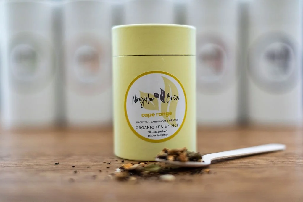 A yellow cylindrical tin of Ningaloo Brew Cape Range organic tea and spice with loose tea leaves and a teaspoon in front of it on a wooden surface, with blurred containers in the background.