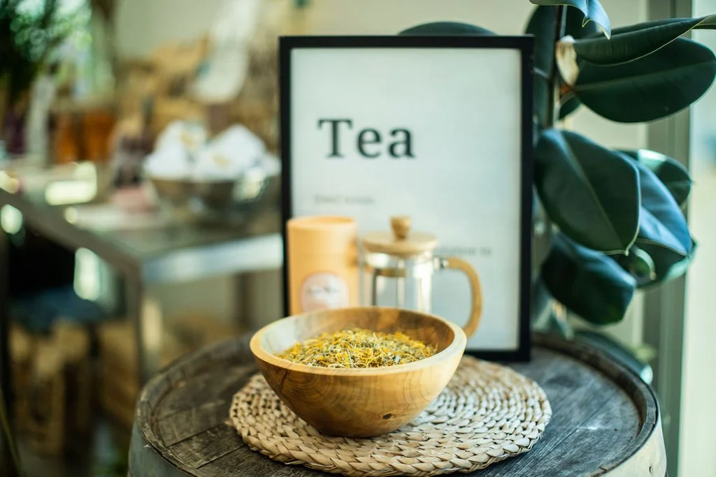 A wooden bowl filled with a dried herbal tea blend on a woven placemat, with a framed sign reading 'Tea' and a ceramic teapot in the background on a rustic table, next to a leafy plant.