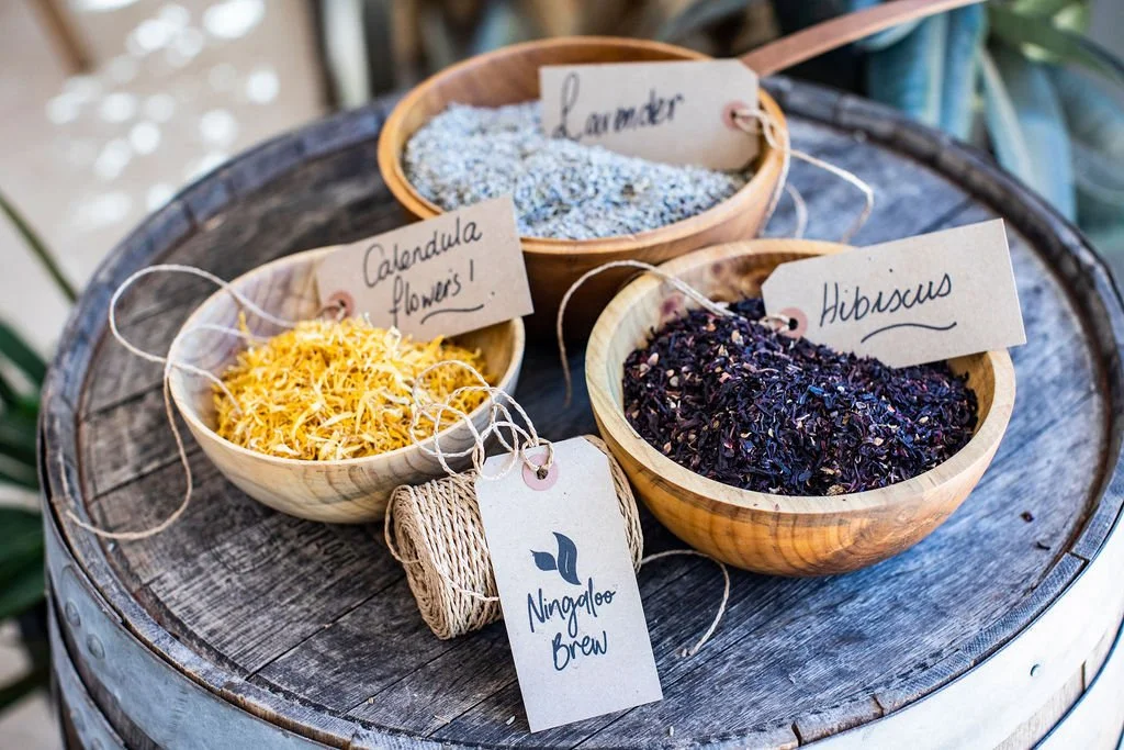 Three wooden bowls containing different dried herbs and flowers. The herbs are labeled as lavender, calendula flowers, and hibiscus. A tag indicates the herbs are for making Ningaloo brew, and a bundle of twine is also on the table.