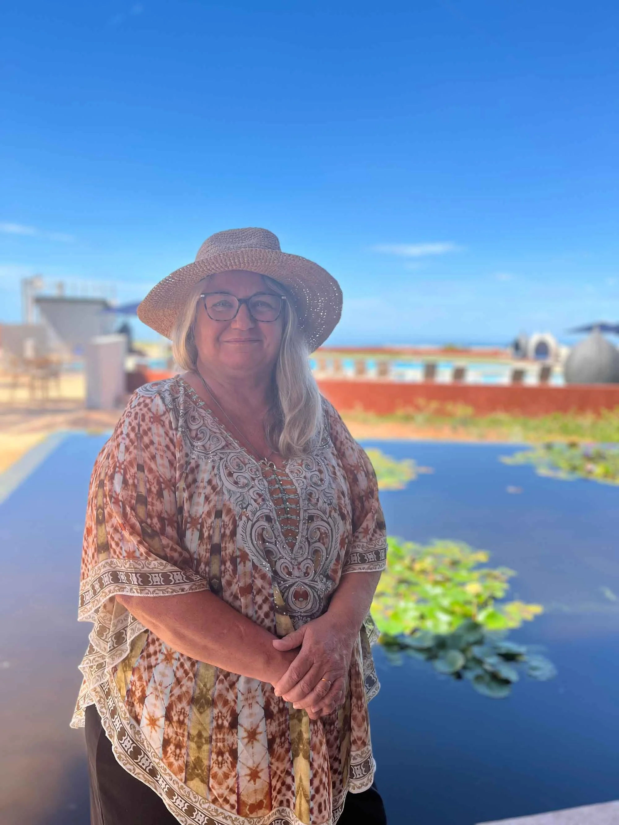 Woman wearing a sunhat and glasses, smiling, standing by a pond with lily pads, with a bright blue sky and ocean in the background.