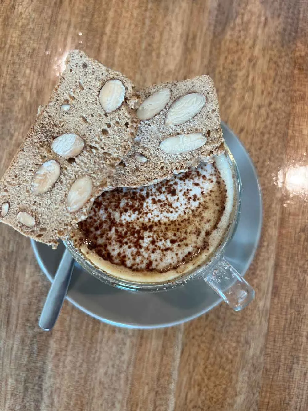 A clear glass cup of coffee topped with foam and sprinkled cocoa powder, served on a gray saucer with a teaspoon, garnished with two almond cookies on top, all on a wooden table.