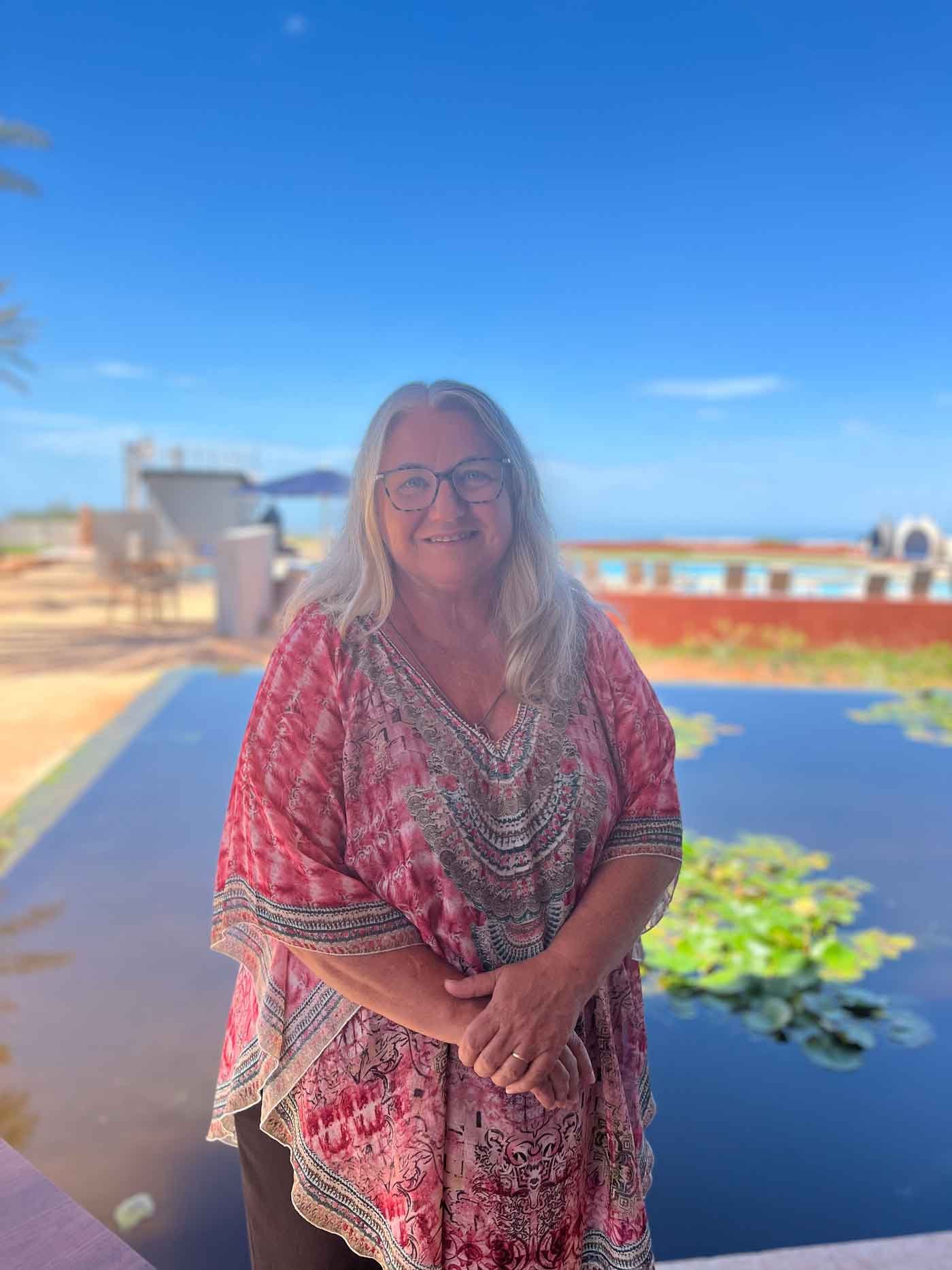 A woman named Darlene with long hair wearing glasses and a red patterned blouse stands by a pond with lily pads, with a clear blue sky and outdoor furniture in the background.