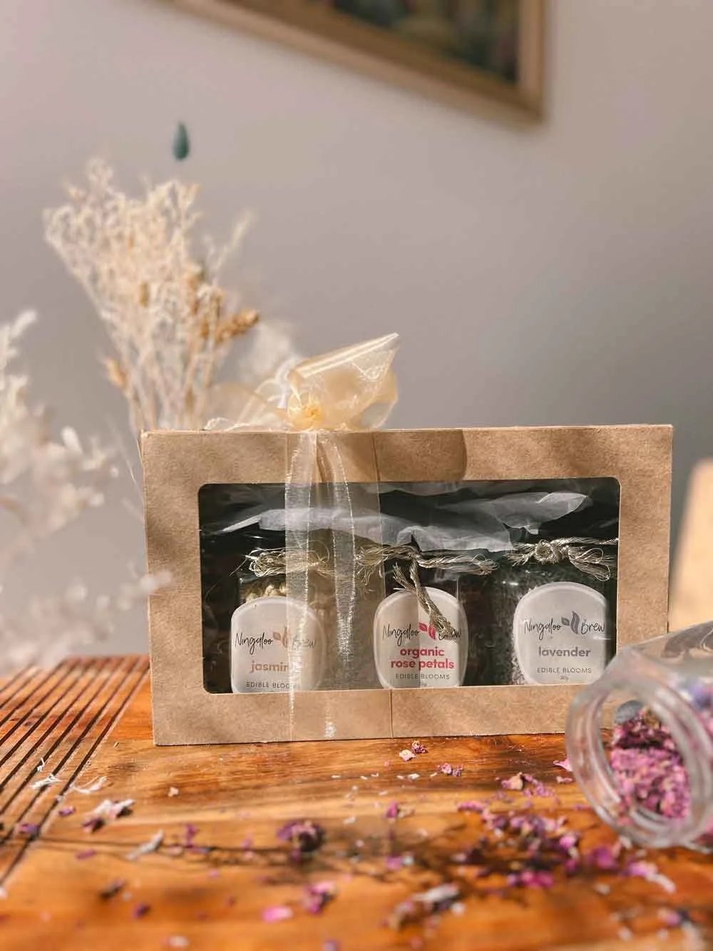 A gift box with three small jars of organic flower petals on a wooden surface, with a jar of dried flower petals spilled nearby and dried flowers in the background.