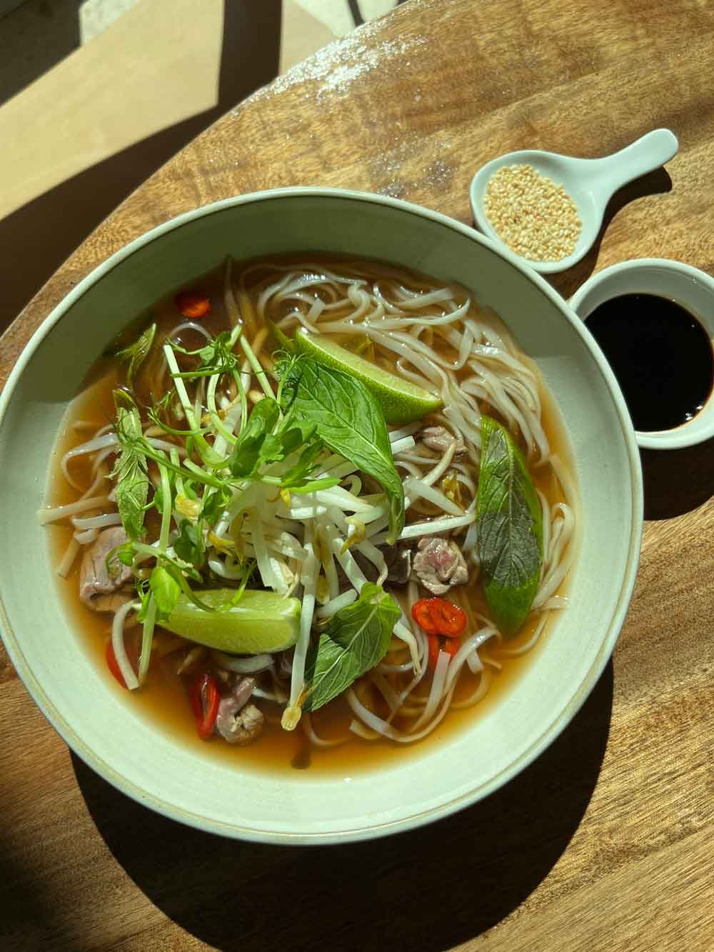 Bowl of Asian noodle soup with vegetables, lime, and herbs, served with soy sauce and sesame seeds on a wooden table.