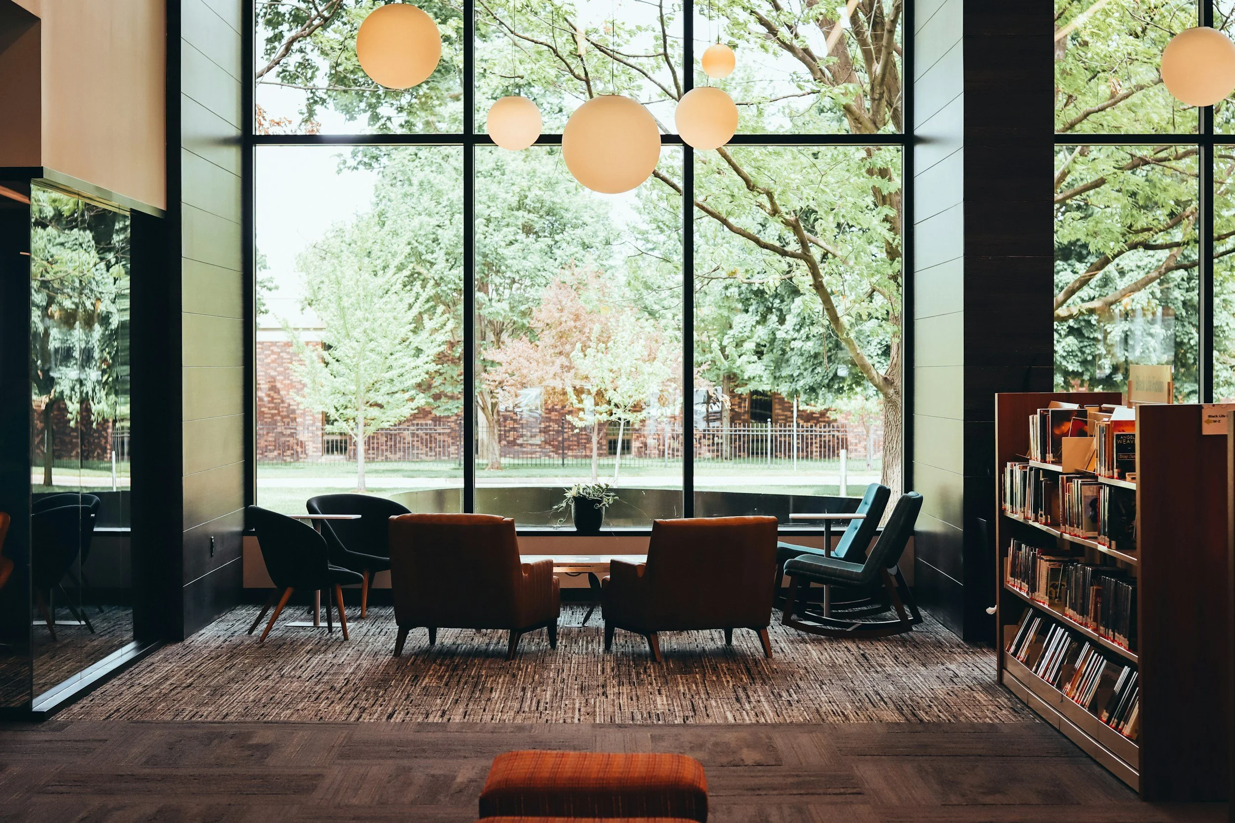 Interior of a room with large floor-to-ceiling windows revealing trees and a grassy area outside. Nearby are black and orange chairs, a wooden bookshelf filled with books, and hanging spherical lamps.