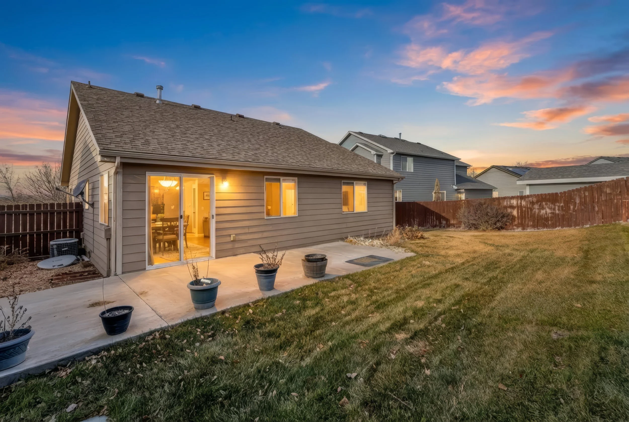 A suburban backyard at sunset with a small gray house, a sliding glass door, potted plants on a concrete patio, and neighboring houses in the background. The sky is colorful with pink and blue hues, and the yard has green grass with some patches of d
