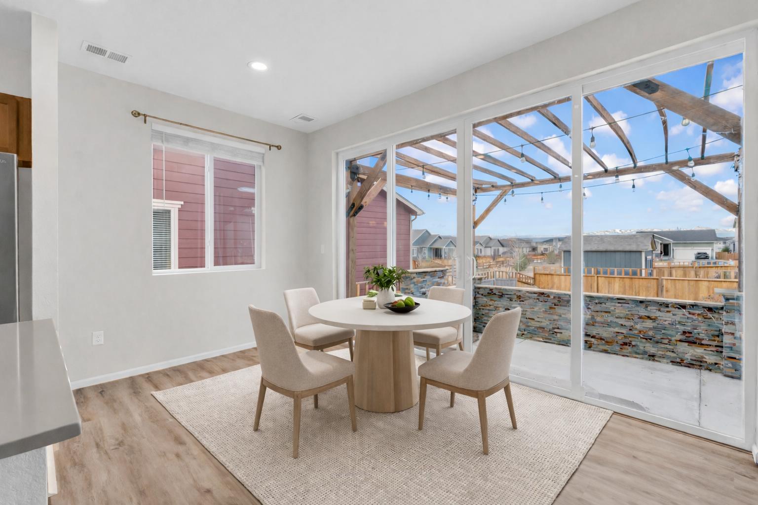 Dining area with a round table, four beige chairs, and a sliding glass door leading to an outdoor patio with a wooden pergola and string lights, in a modern home.