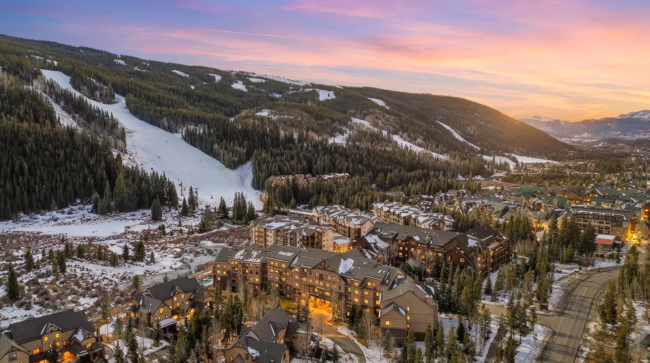 Aerial view of a mountainous town with snow-covered rooftops and winding roads at sunset, surrounded by forested hills and ski slopes.