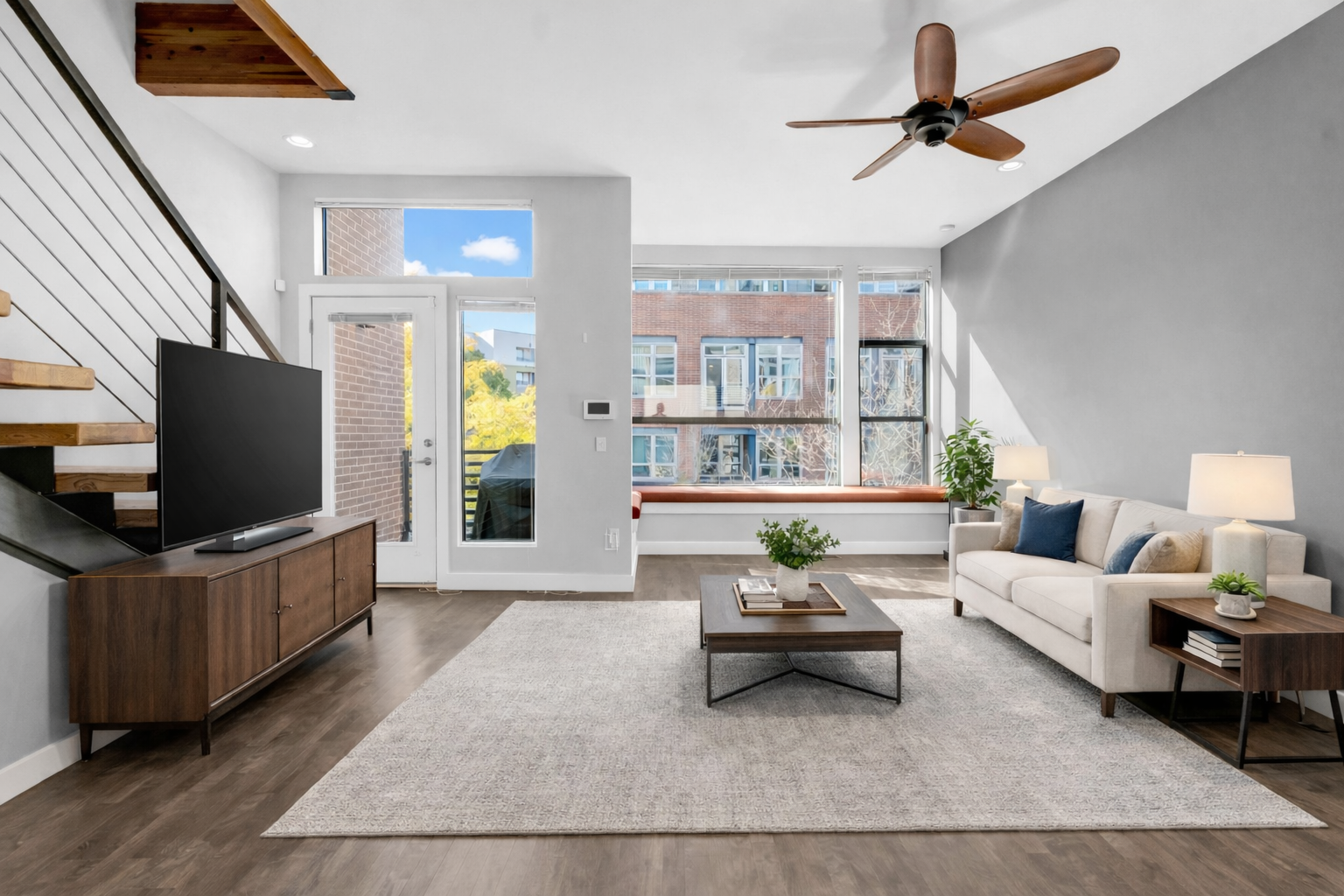 Bright living room with white walls, large windows, a white sofa with cushions, a wooden coffee table, a TV on a wooden stand, and a ceiling fan.