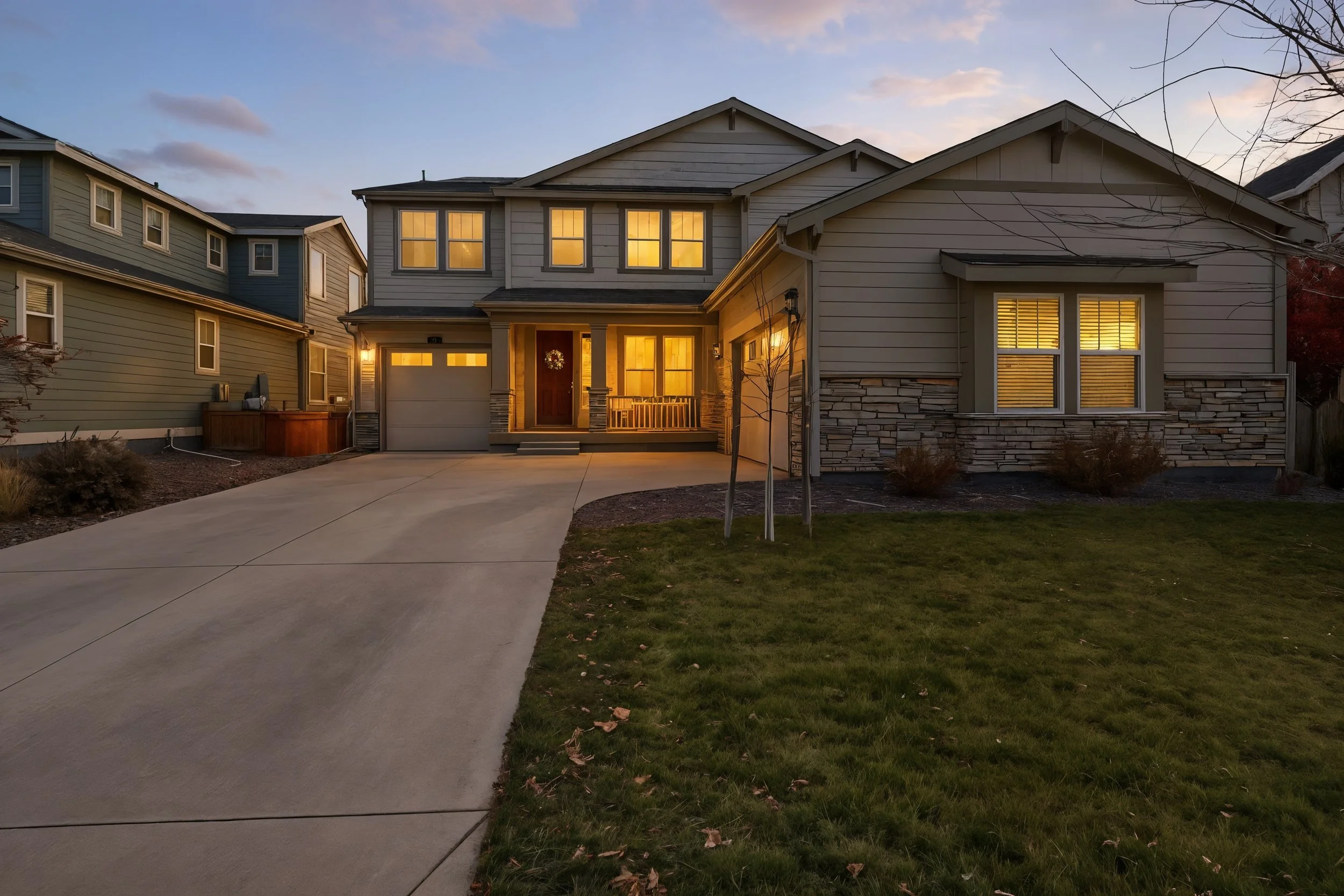 A two-story modern house with front porch and attached garage illuminated by interior lights at dusk, with a concrete driveway and lawn in the foreground.