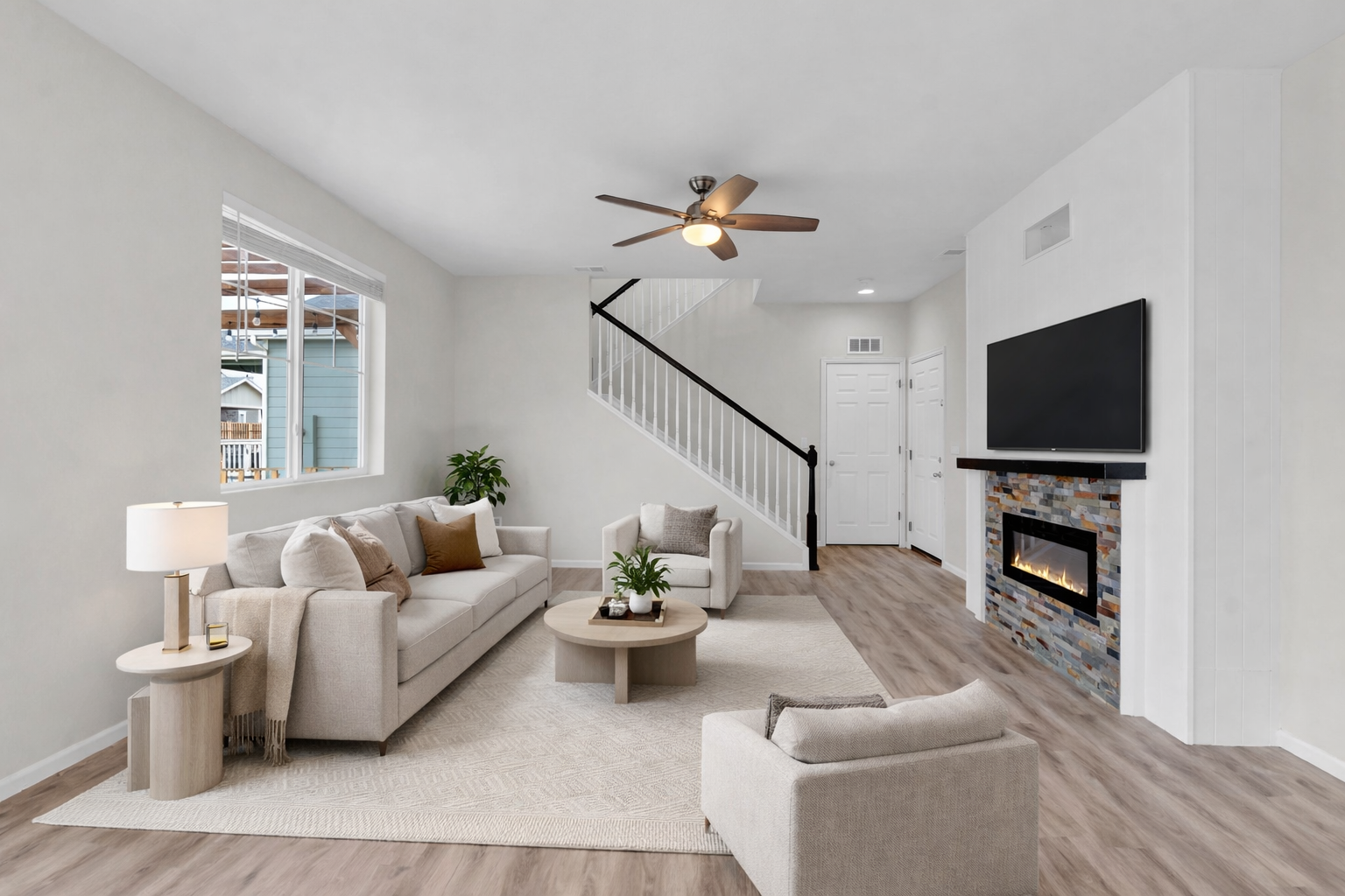 Living room with white walls, beige sofa and chairs, wooden coffee table, fireplace with stone surround, mounted TV, ceiling fan, large window, hardwood floors, and decorative pillows.