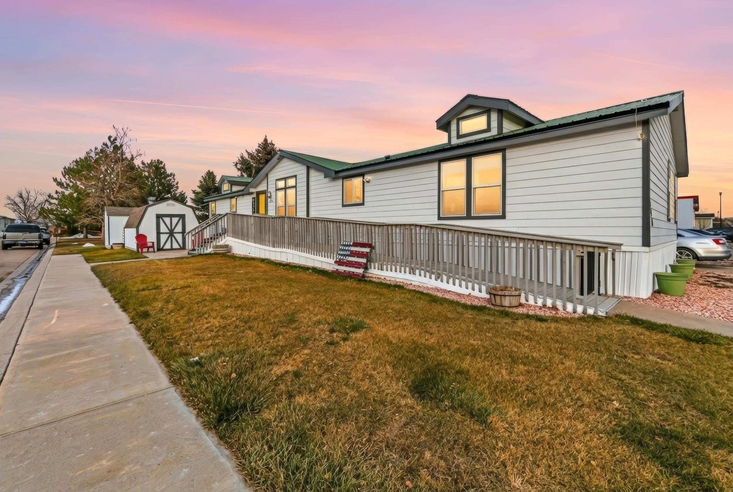 A white mobile home with a green roof, multiple windows, and a wooden ramp leading to the entrance. The yard has green and brown grass, two green planters, and a wooden barrel. There is a sidewalk in front and a gravel driveway with parked cars to the side. The sky is pink and purple during sunset.