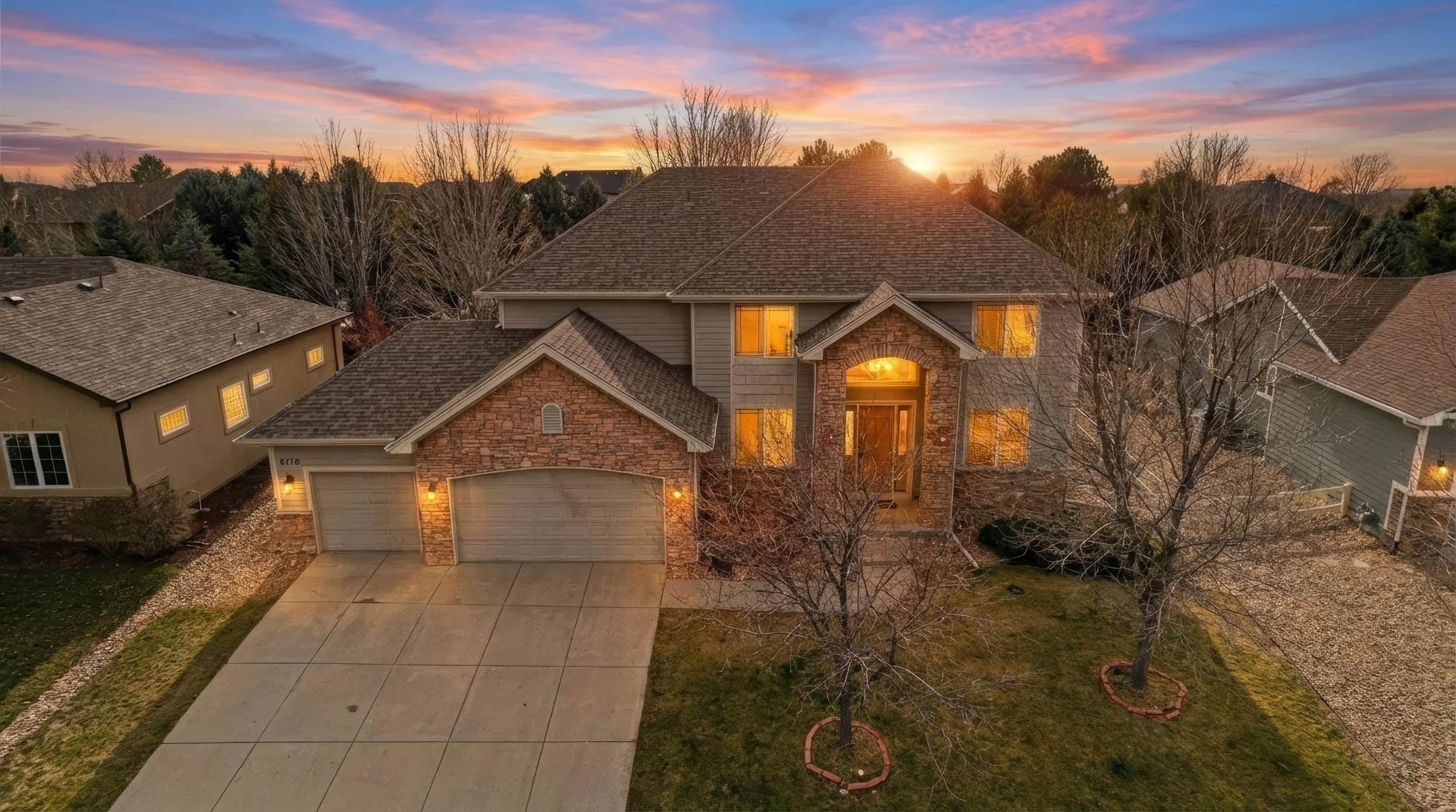 A two-story house with a brick facade and a brown roof, illuminated by exterior lights at sunset, with leafless trees and neighboring houses nearby.
