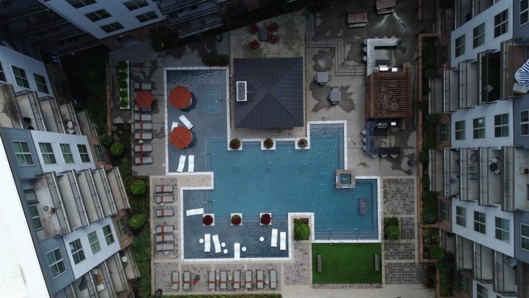 A bird's eye view of a modern apartment complex with a central outdoor pool. Surrounding the pool are lounge chairs, umbrellas, and small landscaped areas with trees and grass. The complex has multiple floors with balconies.