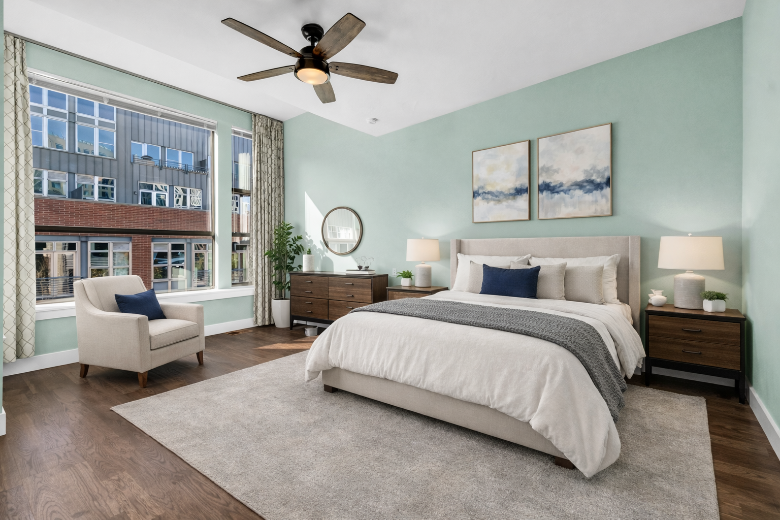 Modern bedroom with large window showcasing city buildings, beige armchair with navy pillow, bed with white and gray bedding, wooden nightstands with lamps, artwork above bed, and a large rug on wooden floor.