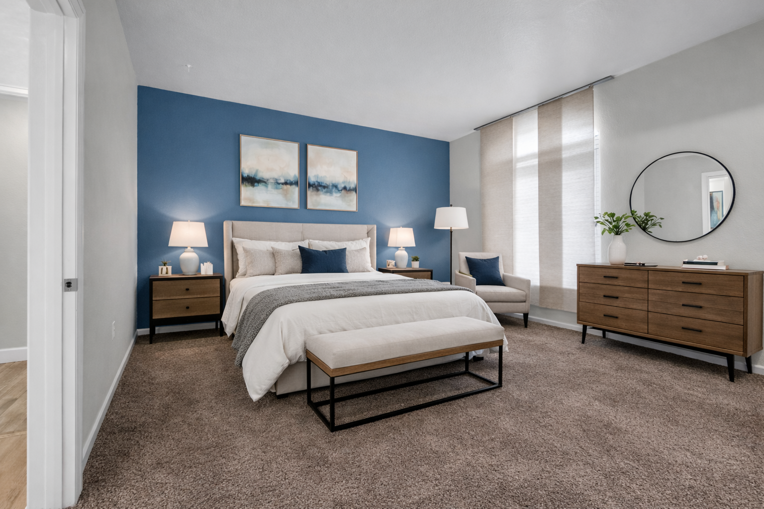 Modern bedroom with beige and blue color scheme, featuring a bed, side tables with lamps, a chair, a dresser, and wall art.