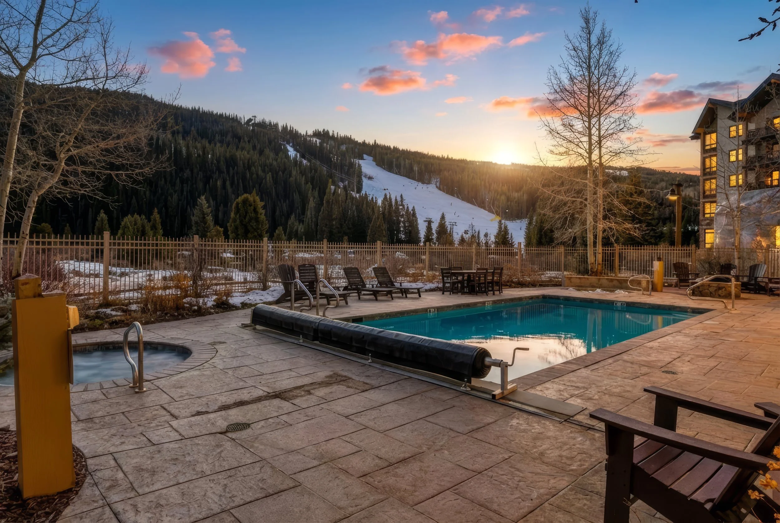 An outdoor pool and hot tub area at sunset, with mountains and a ski slope in the background, surrounded by a wooden fence, leafless trees, and a building with lit windows.