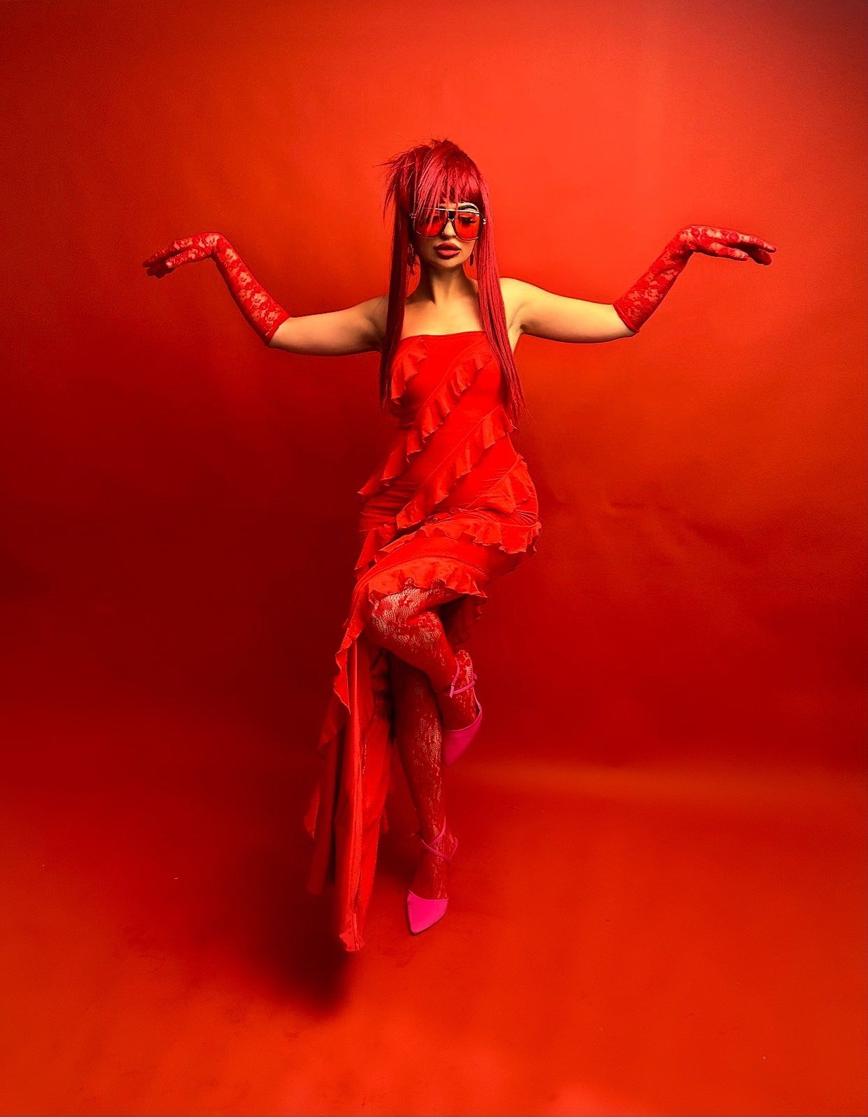 Person in a red dress and sunglasses posing against a red background, wearing long red gloves and pink shoes.
