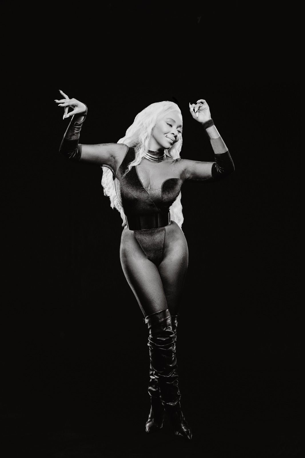 Black and white photo of a person in a form-fitting outfit and knee-high boots dancing against a dark background.