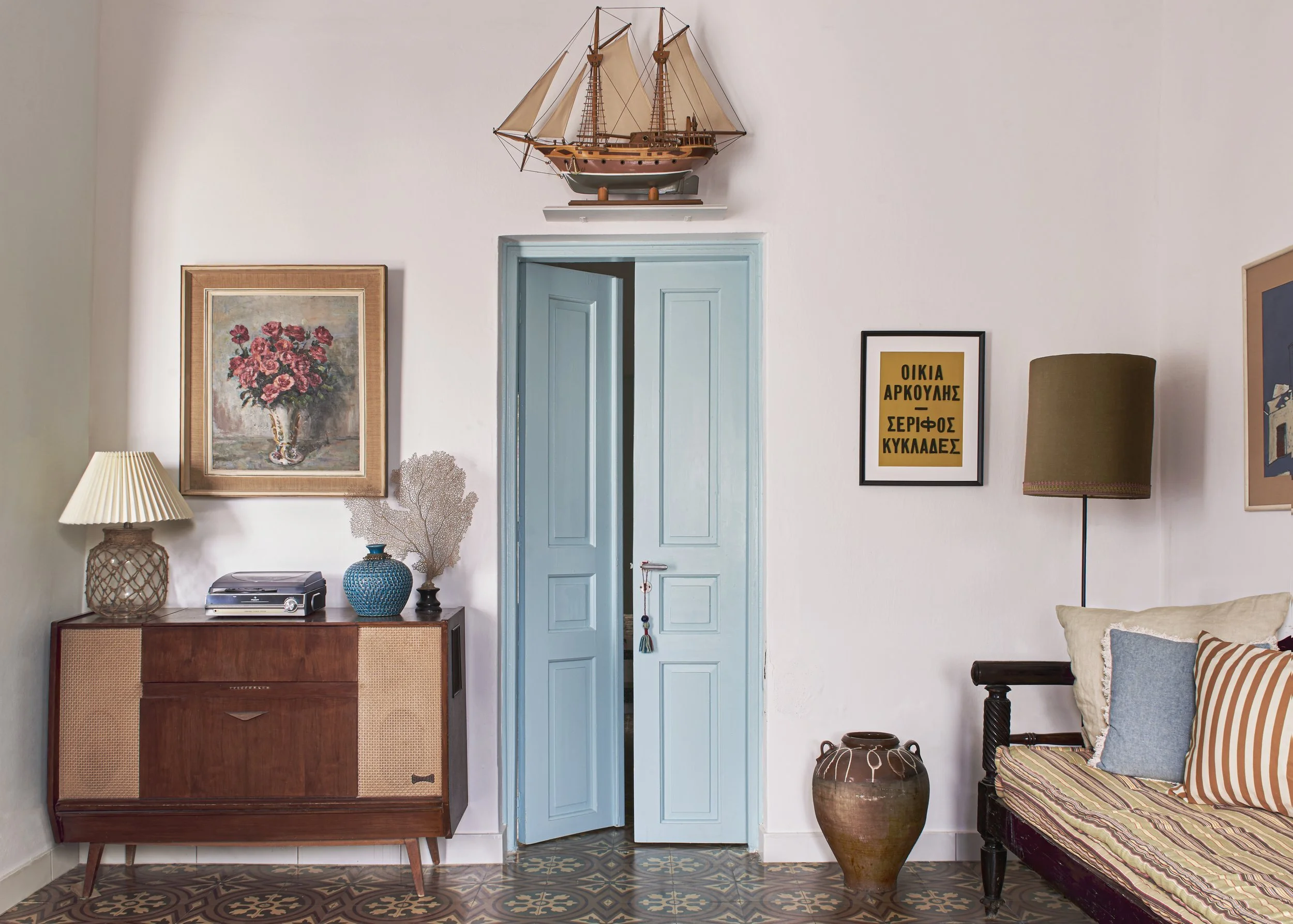 1950s Greek island hallway featuring original restored floor tiles, a vintage mid-century sideboard, and pale green double doors.