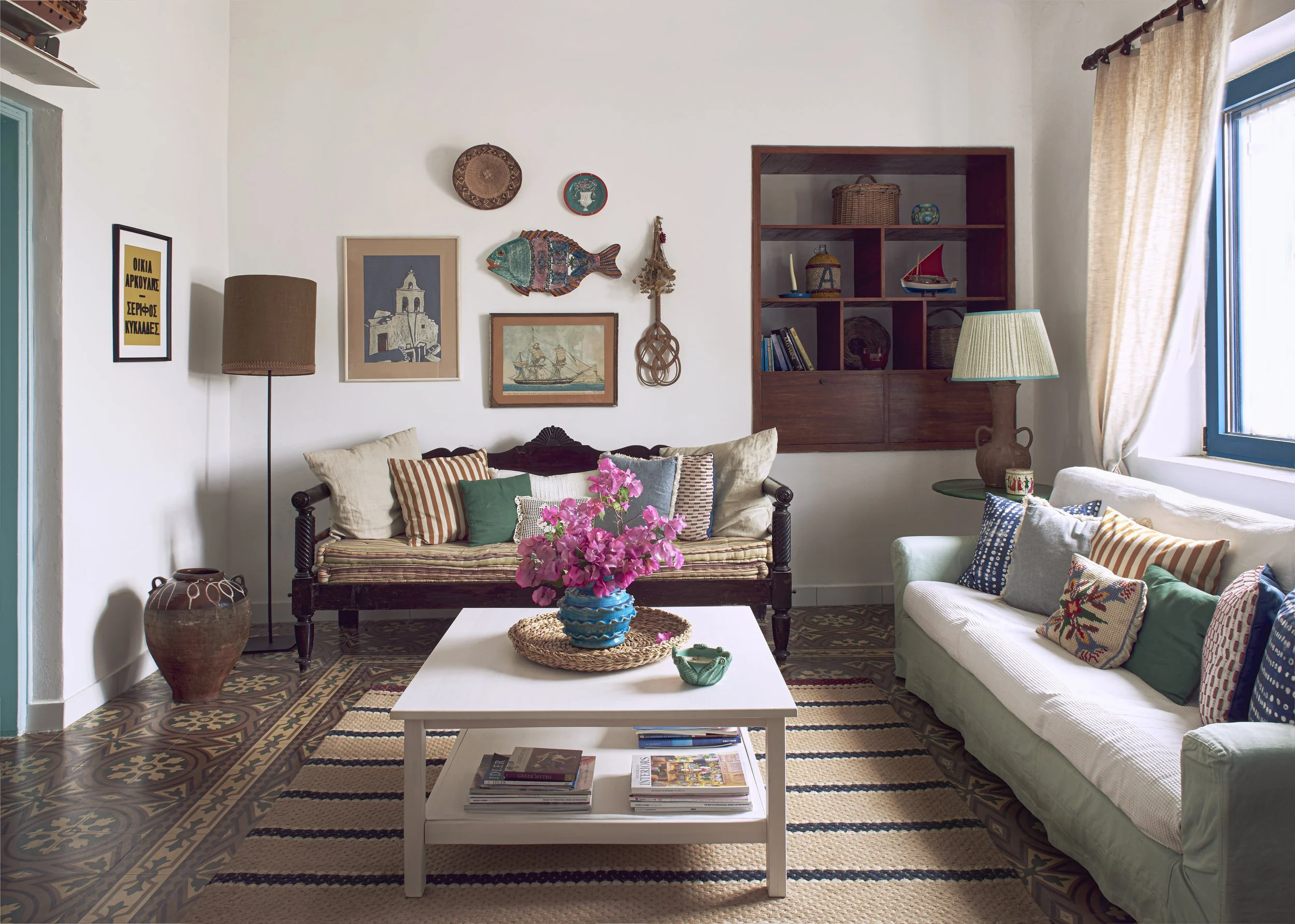 Eclectic Greek island living room interior featuring original 1950s floor tiles, built-in wooden shelving, and colorful vintage cushions.