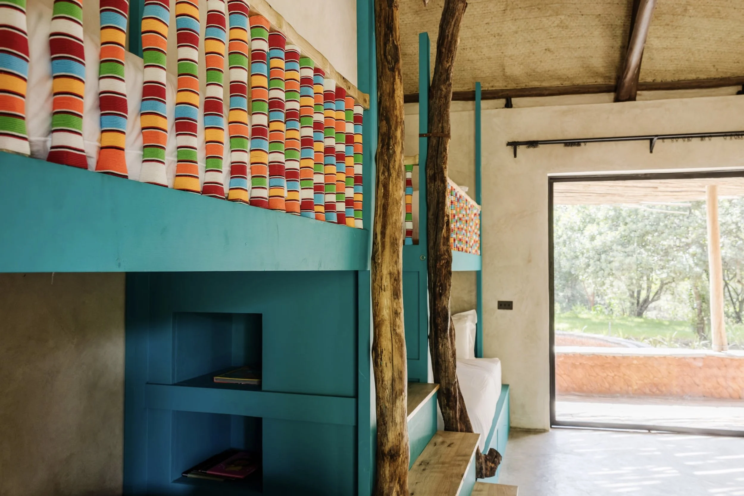 Custom bright blue bunk beds with colorful striped bolsters and natural timber posts in a Kenyan safari lodge by Howark Design.