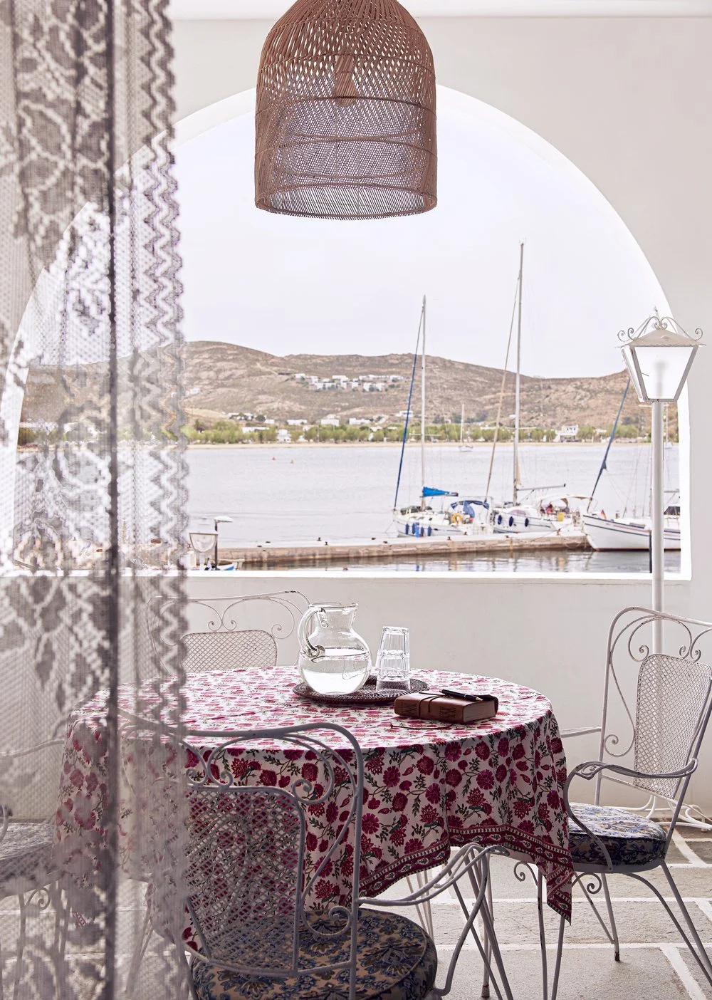 Shaded Greek villa terrace overlooking a harbor, featuring a red floral tablecloth and vintage wire dining chairs.