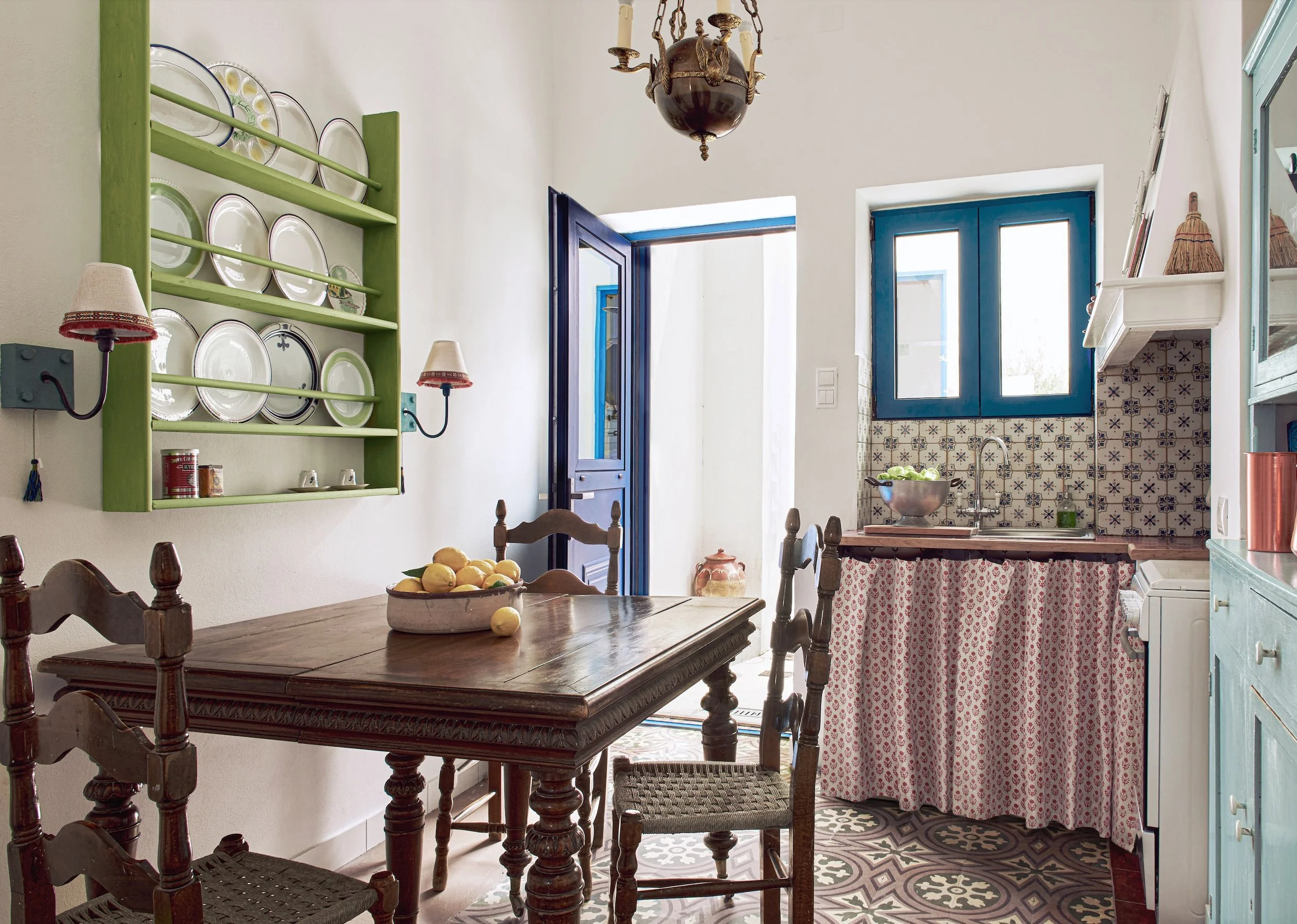 Eclectic Mediterranean kitchen in a Serifos island home featuring patterned floor tiles, blue window frames, and a vintage dining table by Howark Design.