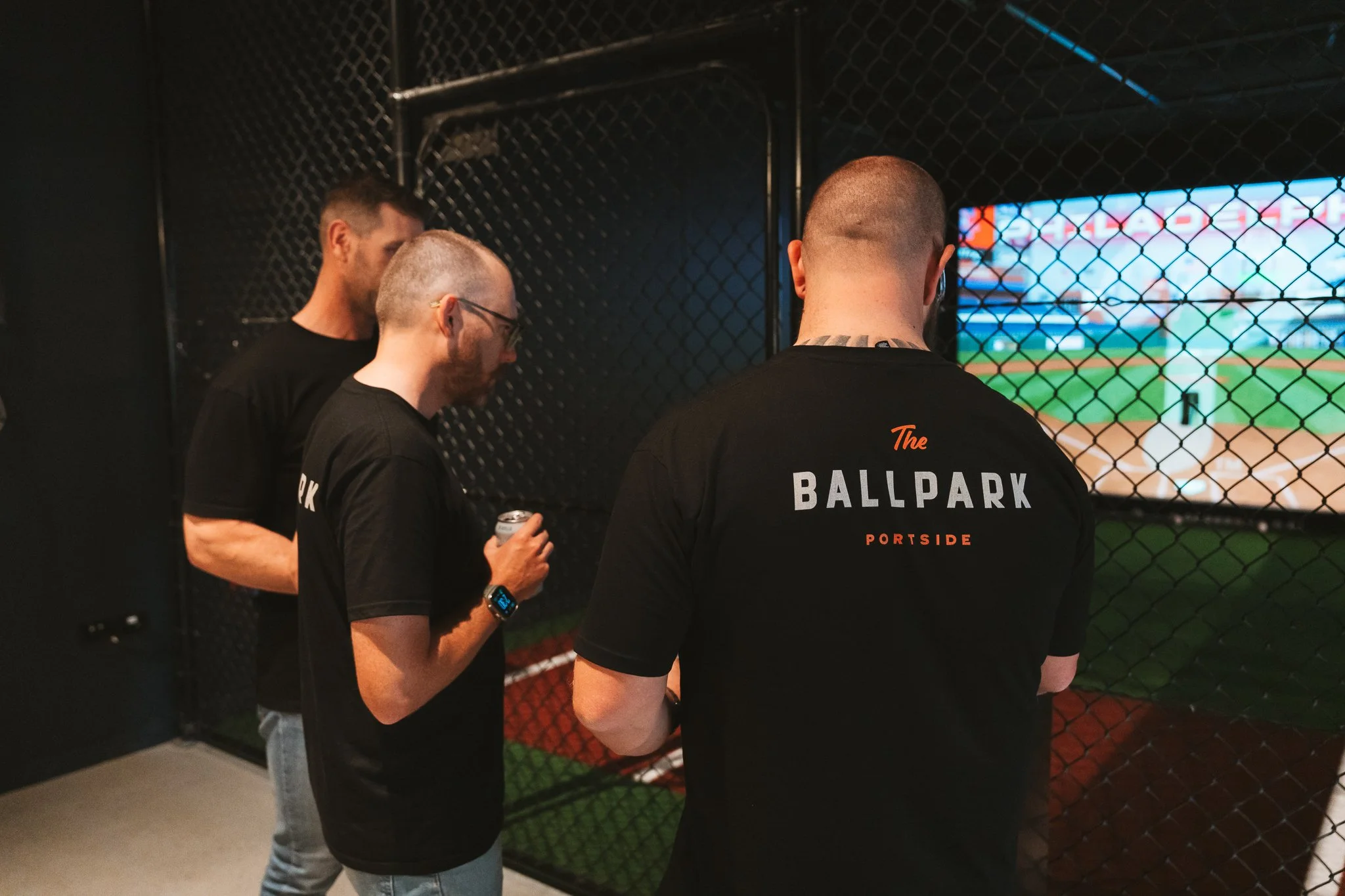 Three men are standing behind a chain-link fence watching a baseball game on a large screen. They are indoors, wearing black shirts with the words "The Ballpark" and "Portside" printed on the back.