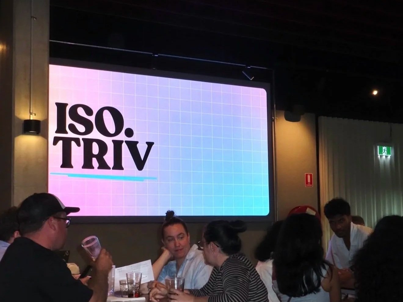 A group of people sitting at a table in a dimly lit room, with a large screen displaying the text 'ISO. TRIV' in bold black letters on a pastel grid background.