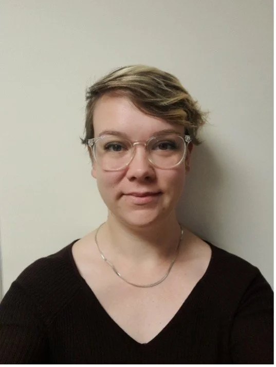 Llun agos o fenyw gyda gwallt golau yn gwenu, yn gwisgo top gwyn, o flaen wal lwyd.
