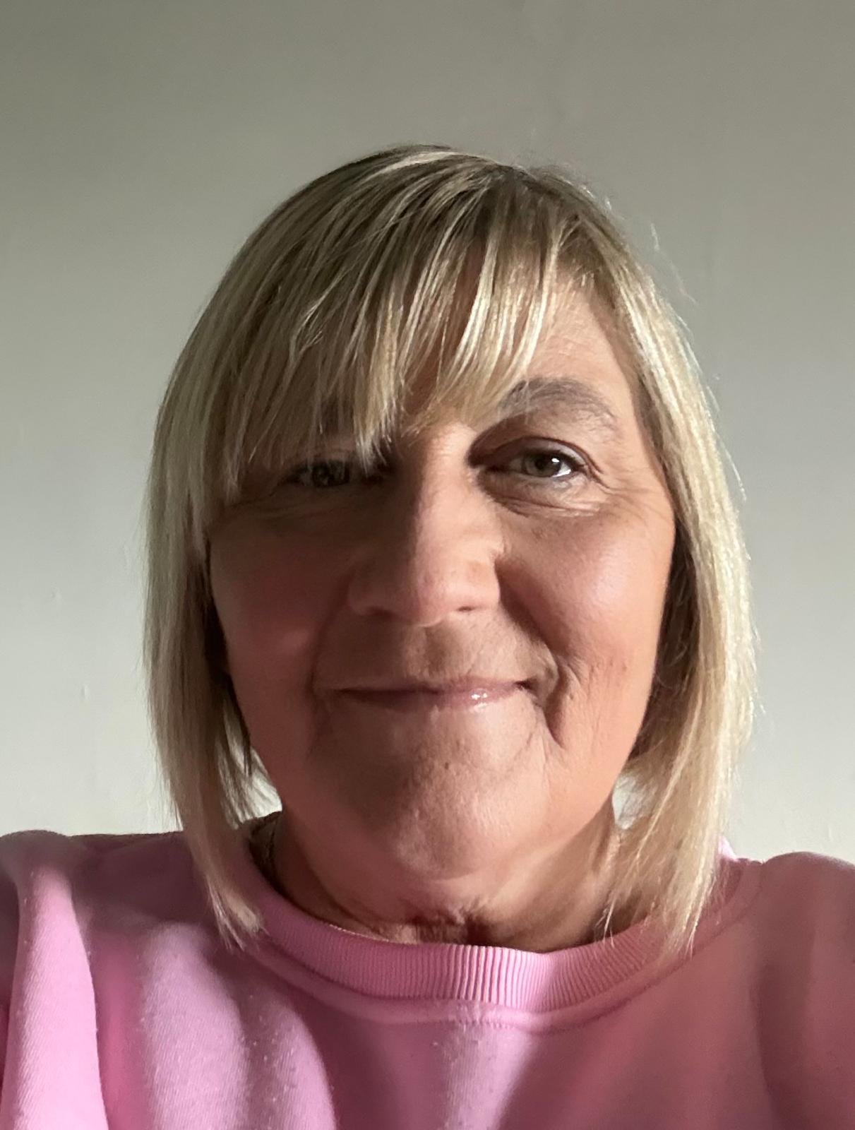 Nyrs gyda gwallt golau a chroen golau yn gwisgo crys pinc, yn gwenu o flaen cefndir plaen.