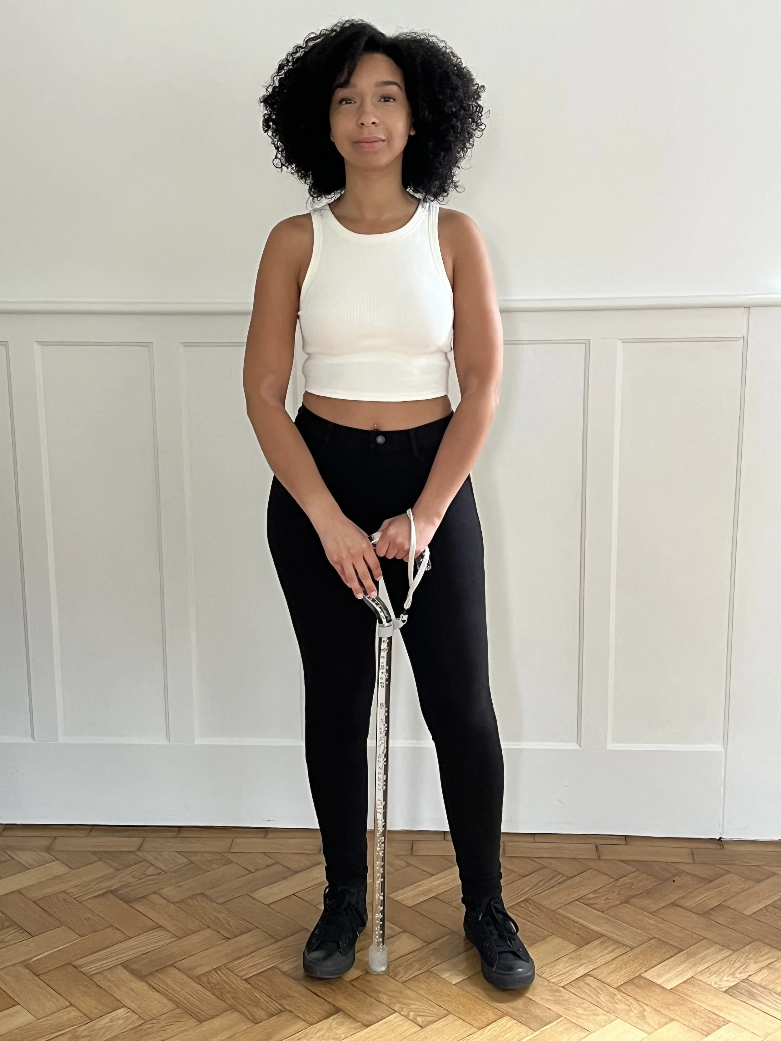 Chloe is stood against a white wall wearing black jeans and a white vest. She’s holding a walking stick in front of her.
