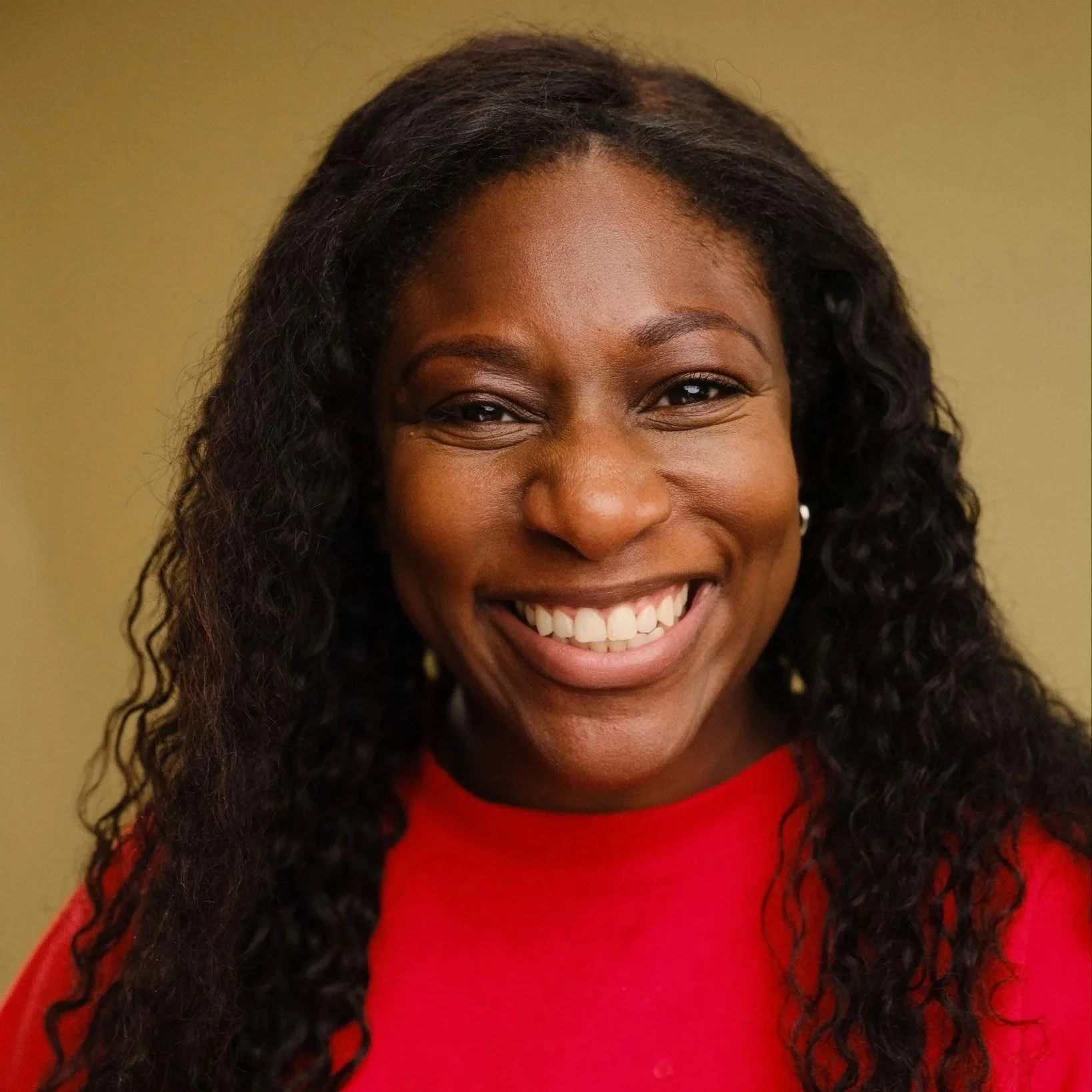 Oneikeh is a Black woman with long curly dark hair. She has brown eyes and is smiling widely, wearing a red top