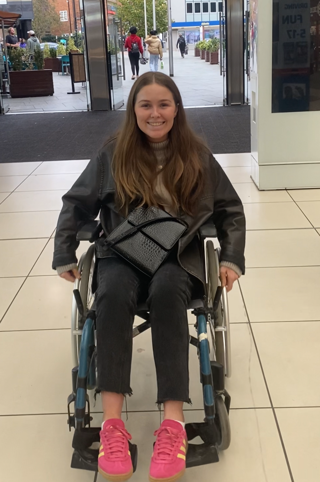 Photo in a shopping centre, sat smiling in a wheelchair, wearing black jeans, a beige jumper and brown leather jacket with bright pink trainers and a black leather handbag on lap.