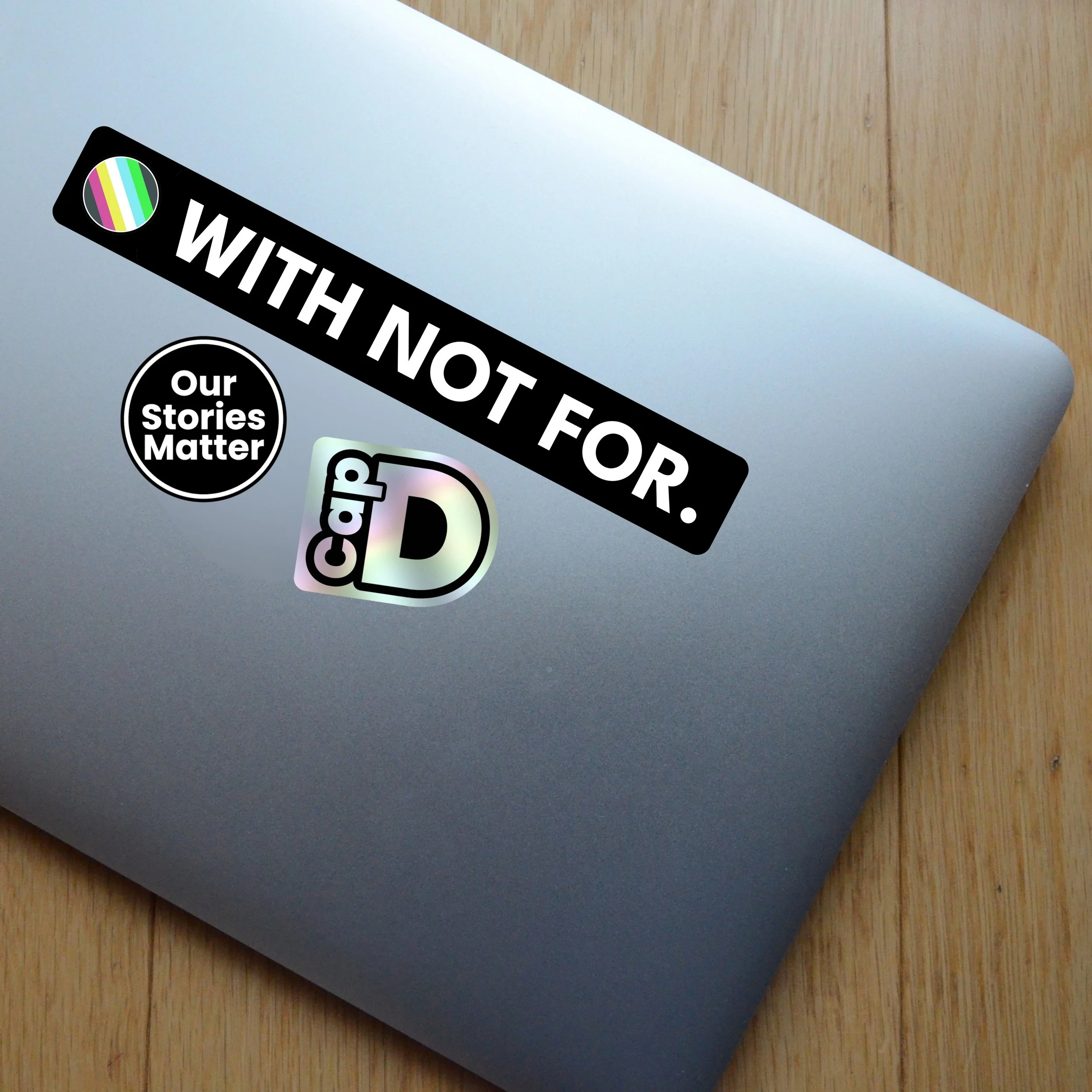 WNF sticker pack on a silver laptop with WITH NOT FOR strip, Our Stories Matter circle and holographic Cap D stickers.jpg.jpg