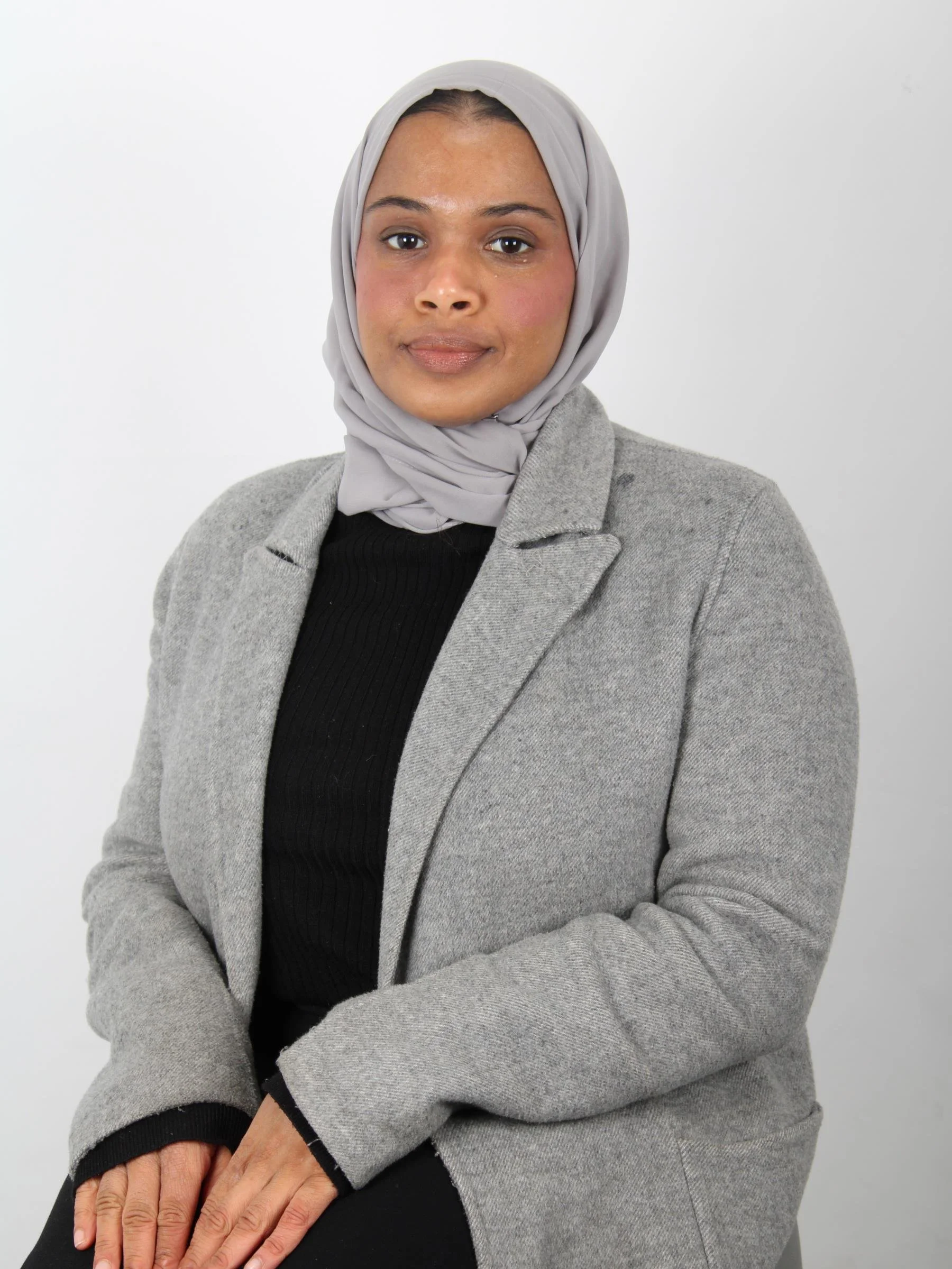 Umi is seated against a plain light grey background, facing the camera with a calm and confident expression. She is wearing a soft grey hijab neatly wrapped around her head, paired with a light grey coat over a black top.