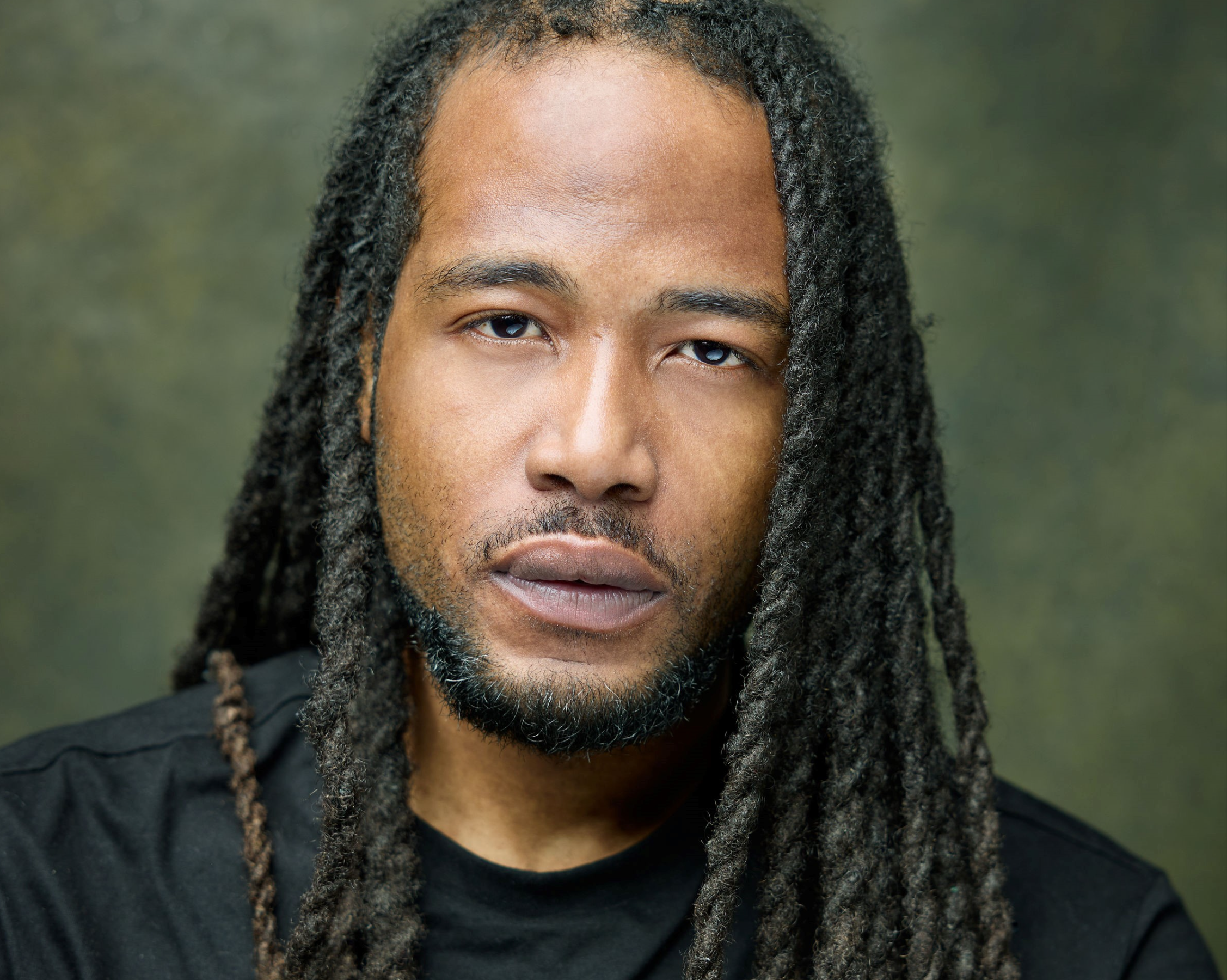 Ekow is a Black man in his 30s with long dark braids and dark facial hair. He is waring a black t.shirt and looking straight at the camera calmly