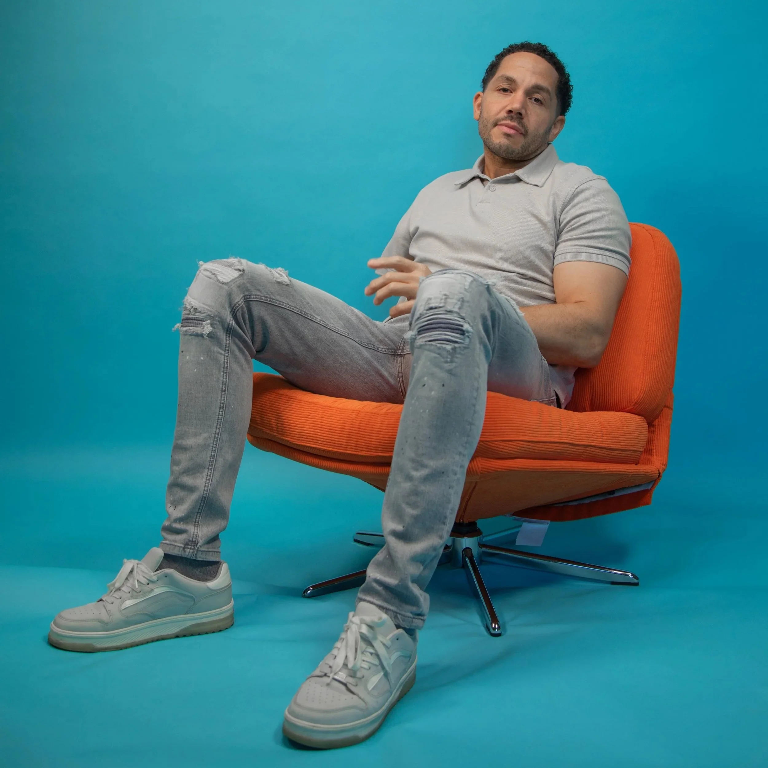 Chris is mixed race with short dark curly hair and brown eyes. He has upper limb difference and sits on an orange chair against a blue background
