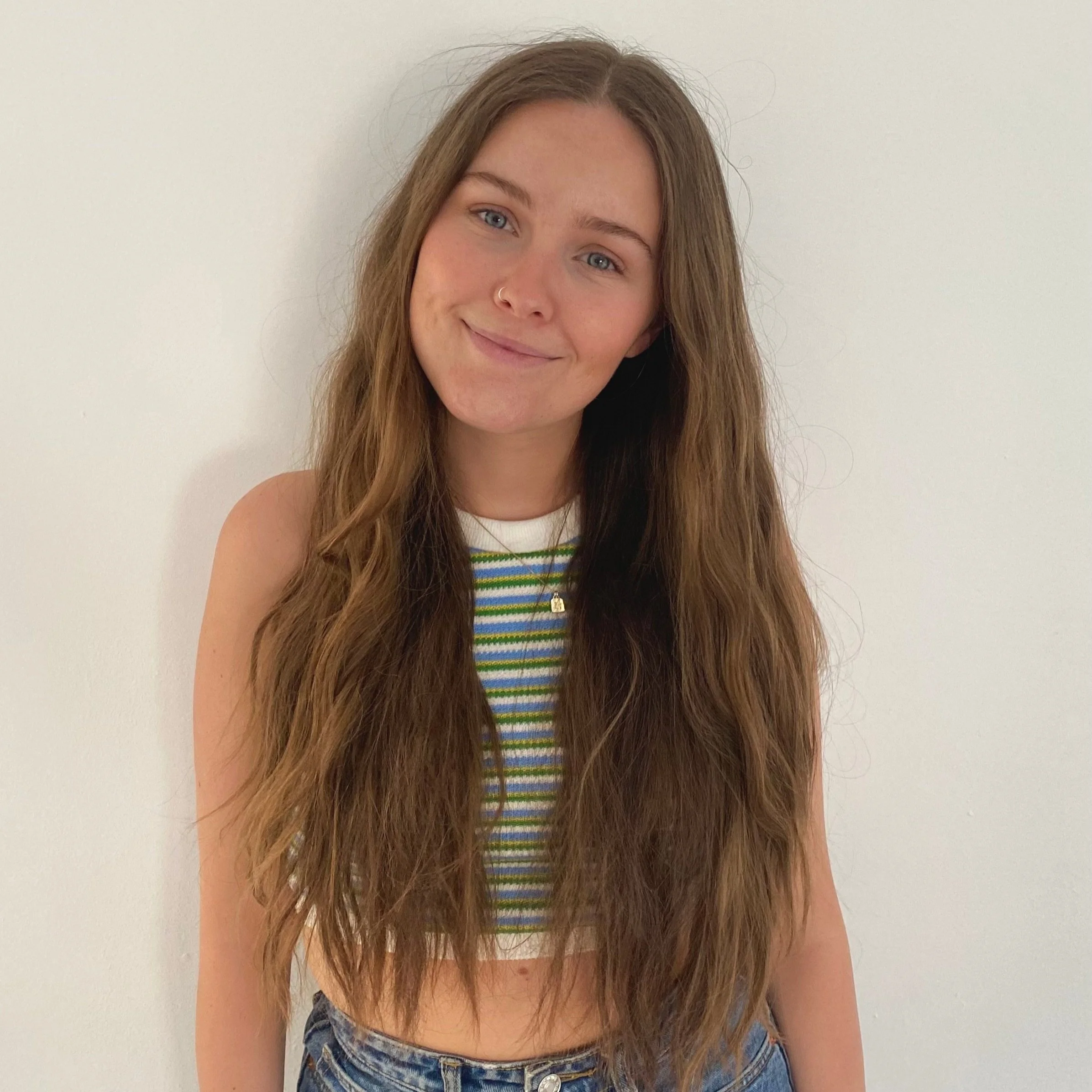 White woman with long light brown hair and blue eyes. Photographed on a plain white background wearing a white, blue, green and yellow horizontal thing striped top.