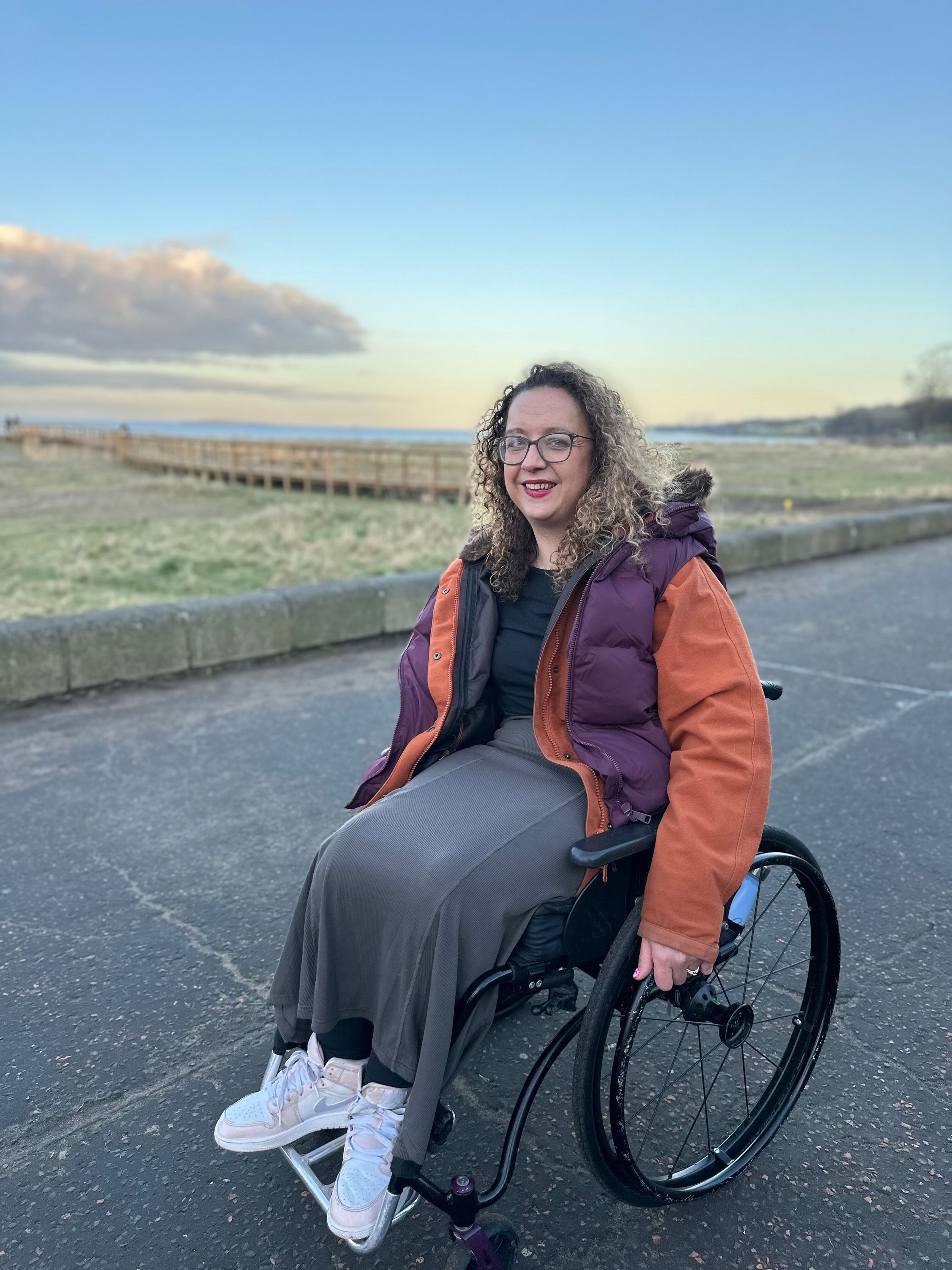 A woman with curly hair wearing glasses and a two-tone winter coat sits in a wheelchair on a path near a coastal boardwalk during sunset. She is smiling and looking at the camera.