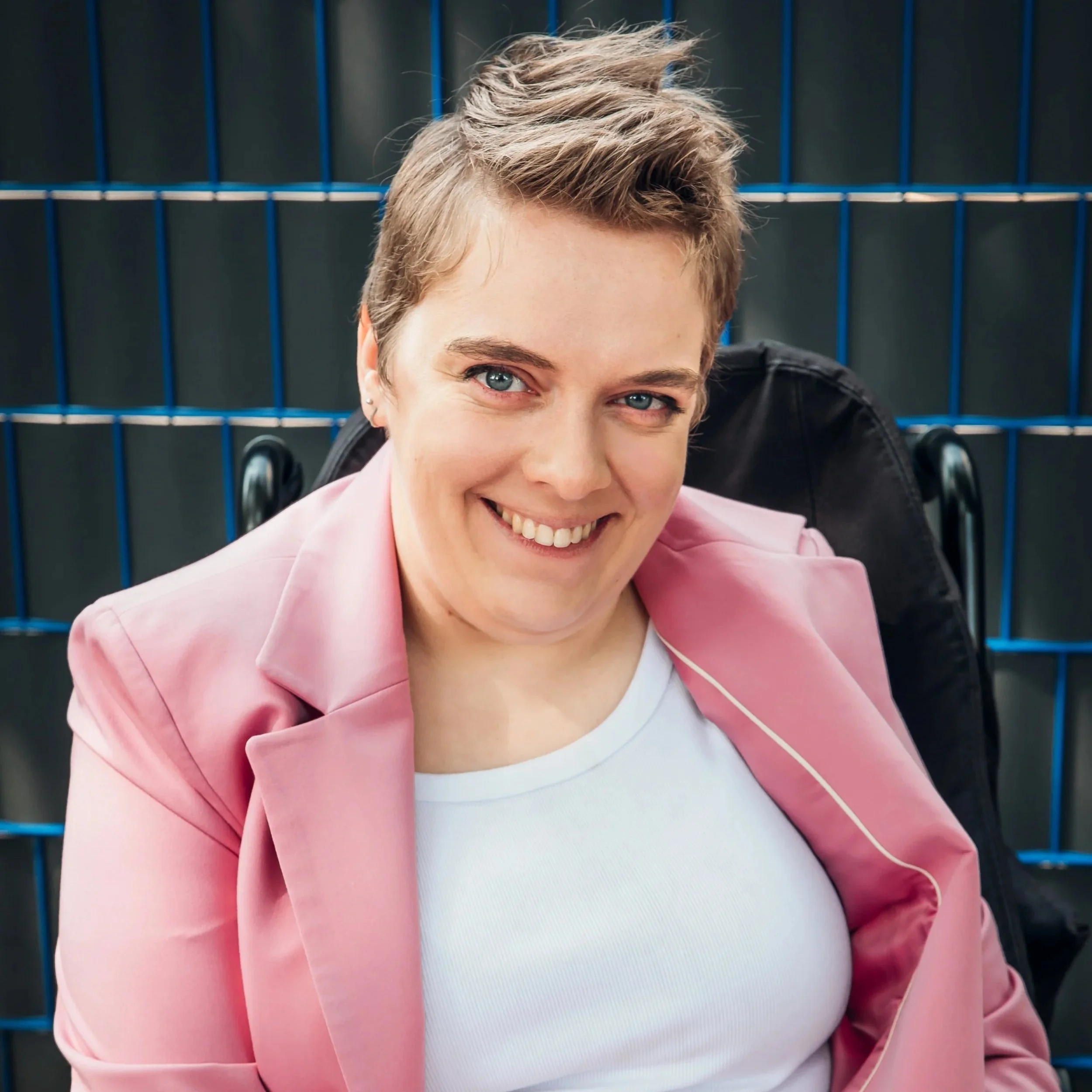 Lucy is a white woman with short brown hair, styled in a quiff at the front. She sits in her wheelchair, wearing a pink suit and white t.shirt.