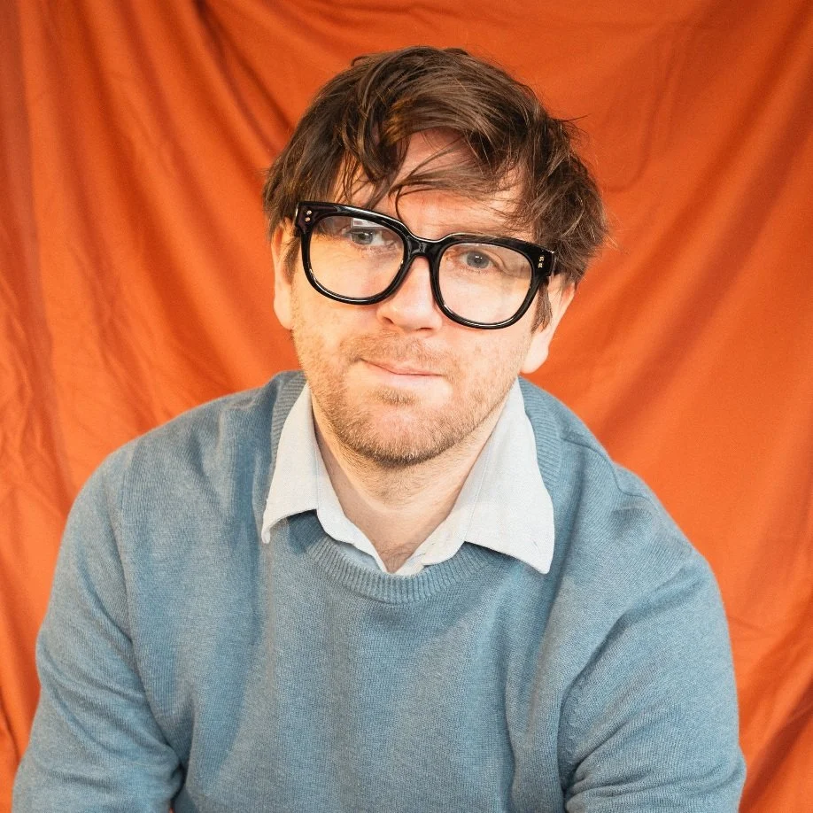 Alexander is a white man with brown hair and stubble. He wears large black rimmed glasses and a grey jumper.
