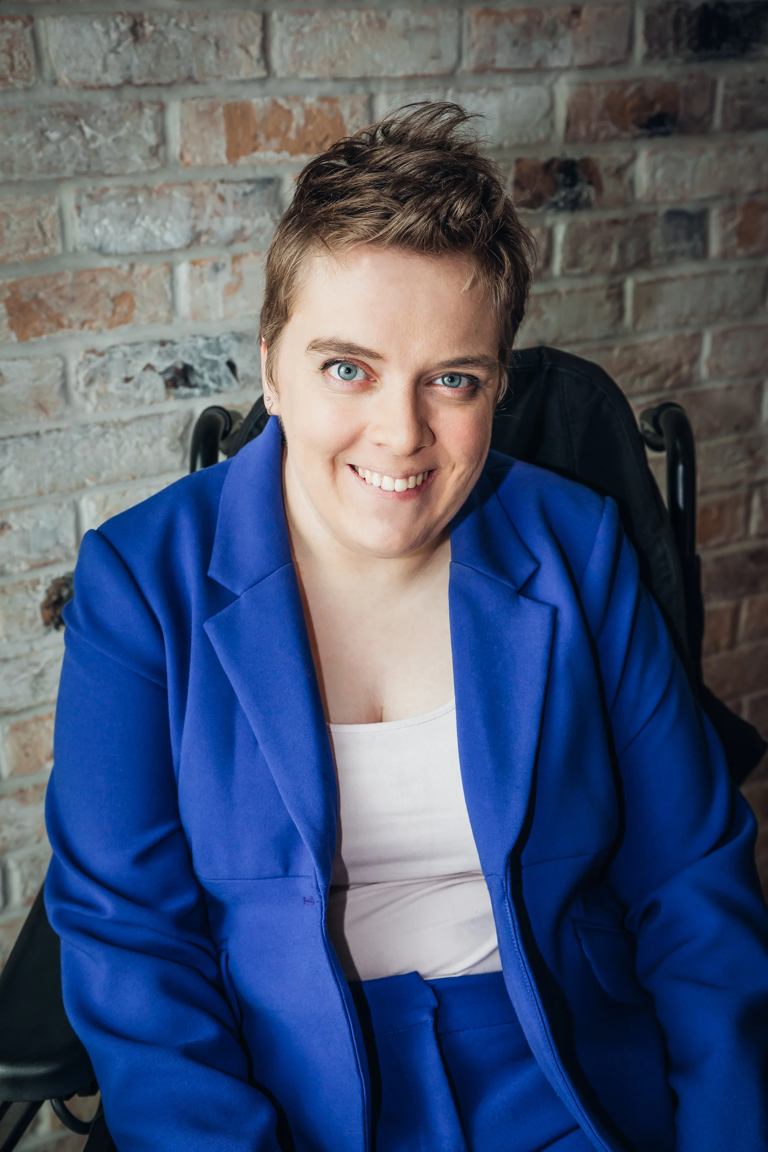 Lucy is sat in her wheelchair in front of a brick wall. She is wearing an electric blue suit with a white vest as she smiles directly into the camera.