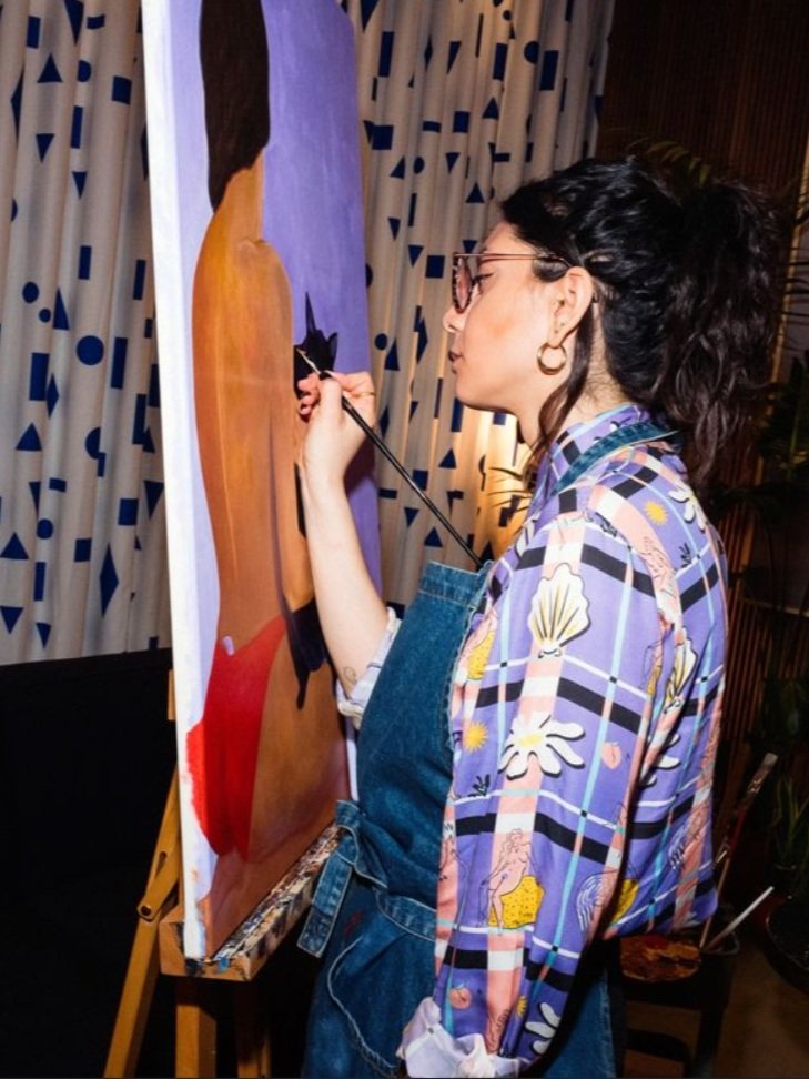 Myriam is painting one of her pieces of the back of a woman in red bikini bottoms on a canvas set on an easel.