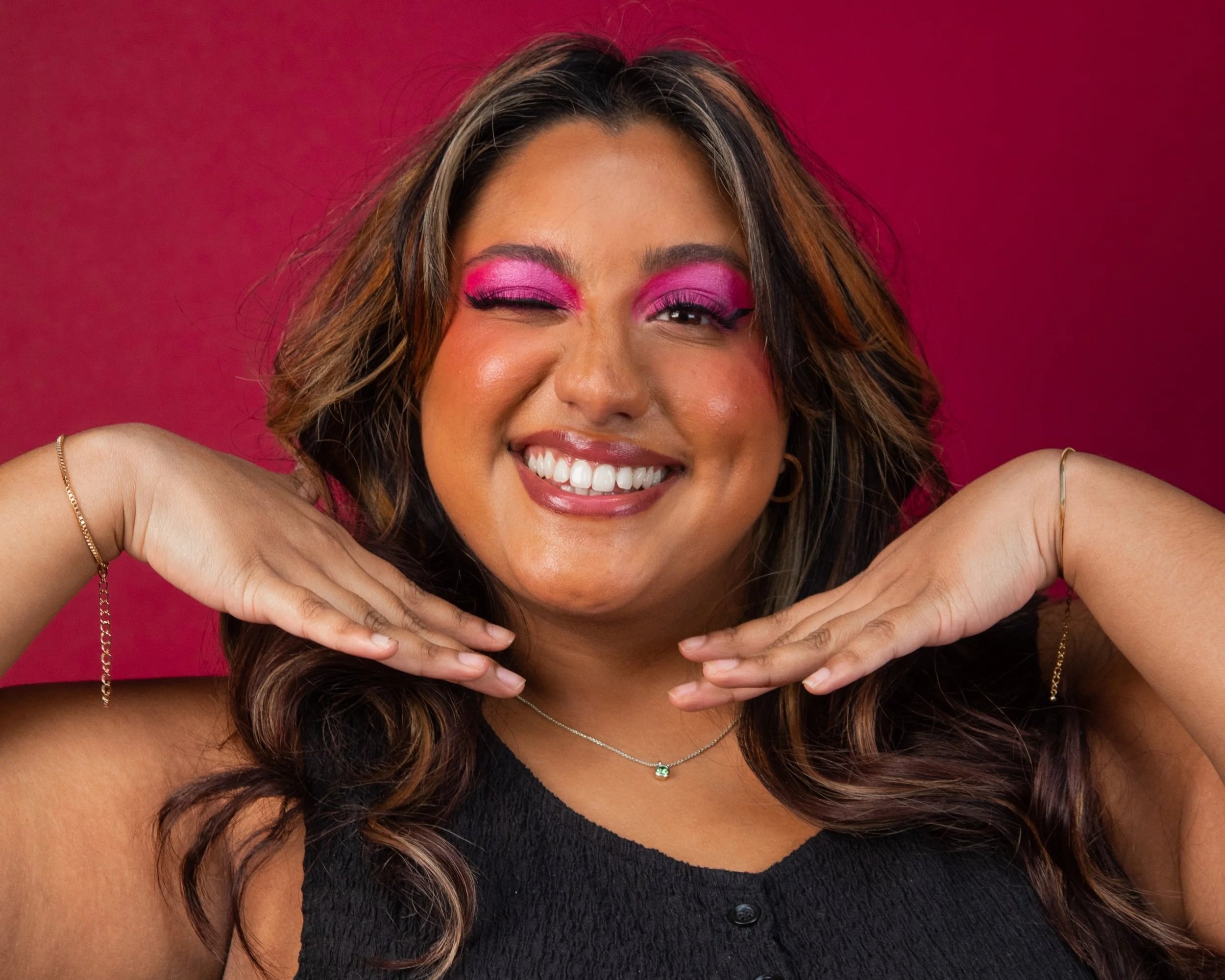 Shareena is a brown woman with long dark curly hair. Her eyeshadow is bright pink as she winks at the camera with her hands placed under her chin. She wears a black vest and silver necklace and is stood in front of a red background.
