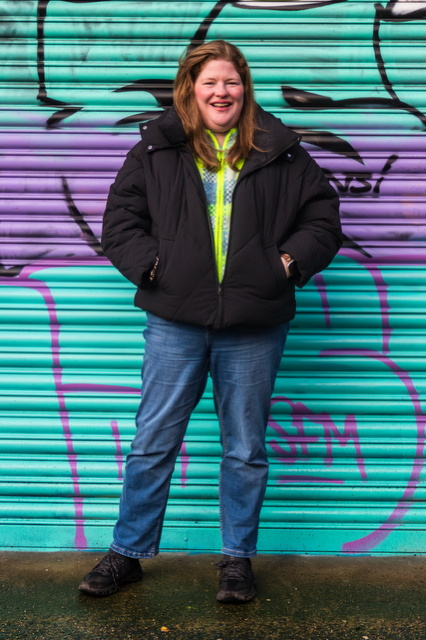 A full-length colour photo of Judi standing in front of a teal and purple graffiti shutter. She is wearing a black puffer jacket and jeans, hands in pockets, smiling broadly.