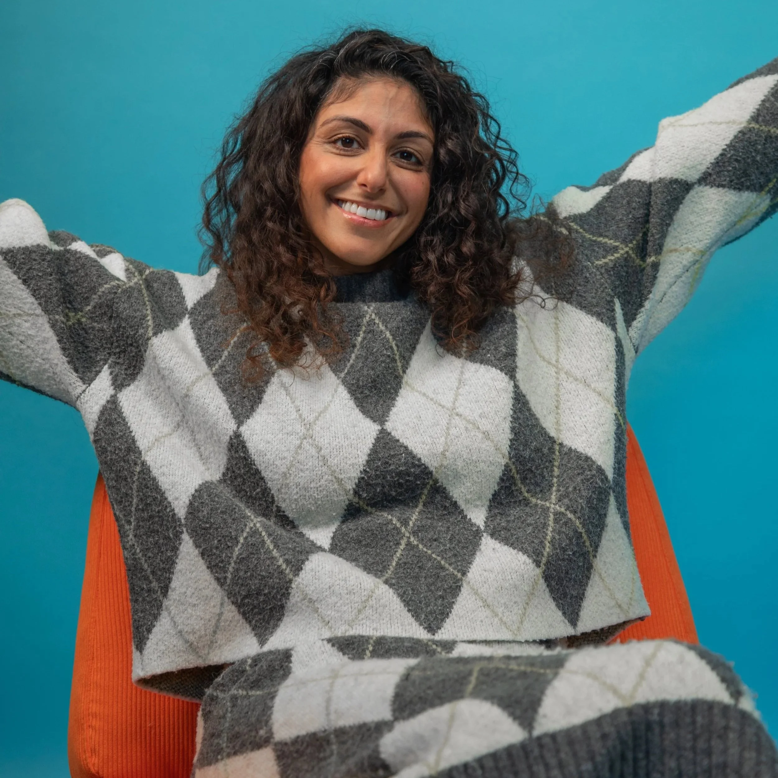 Amanprit is sitting on an orange chair with her arms stretched out to the sides.