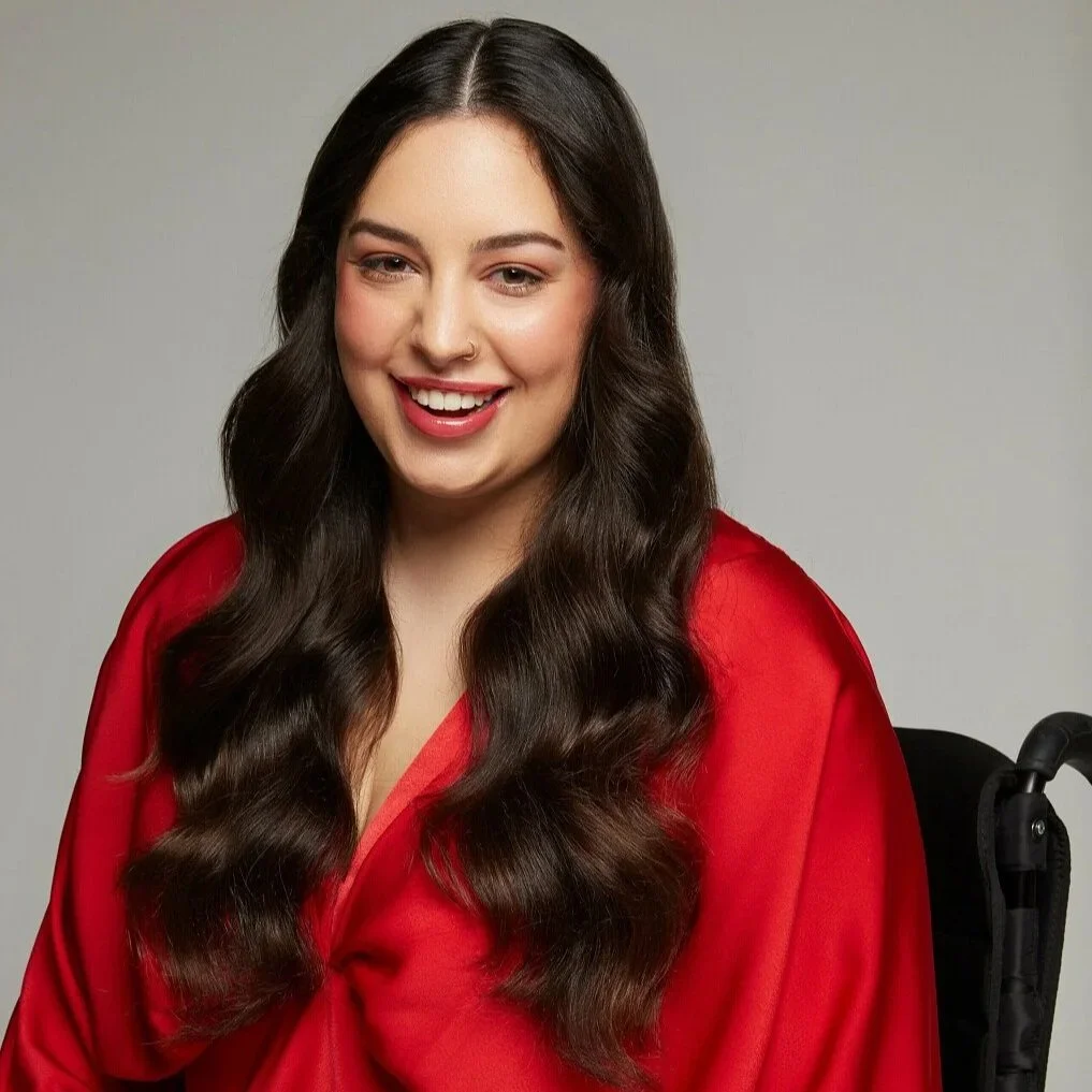 A portrait of Roisin sitting in a wheelchair and smiling brightly, wearing a vibrant red satin dress.