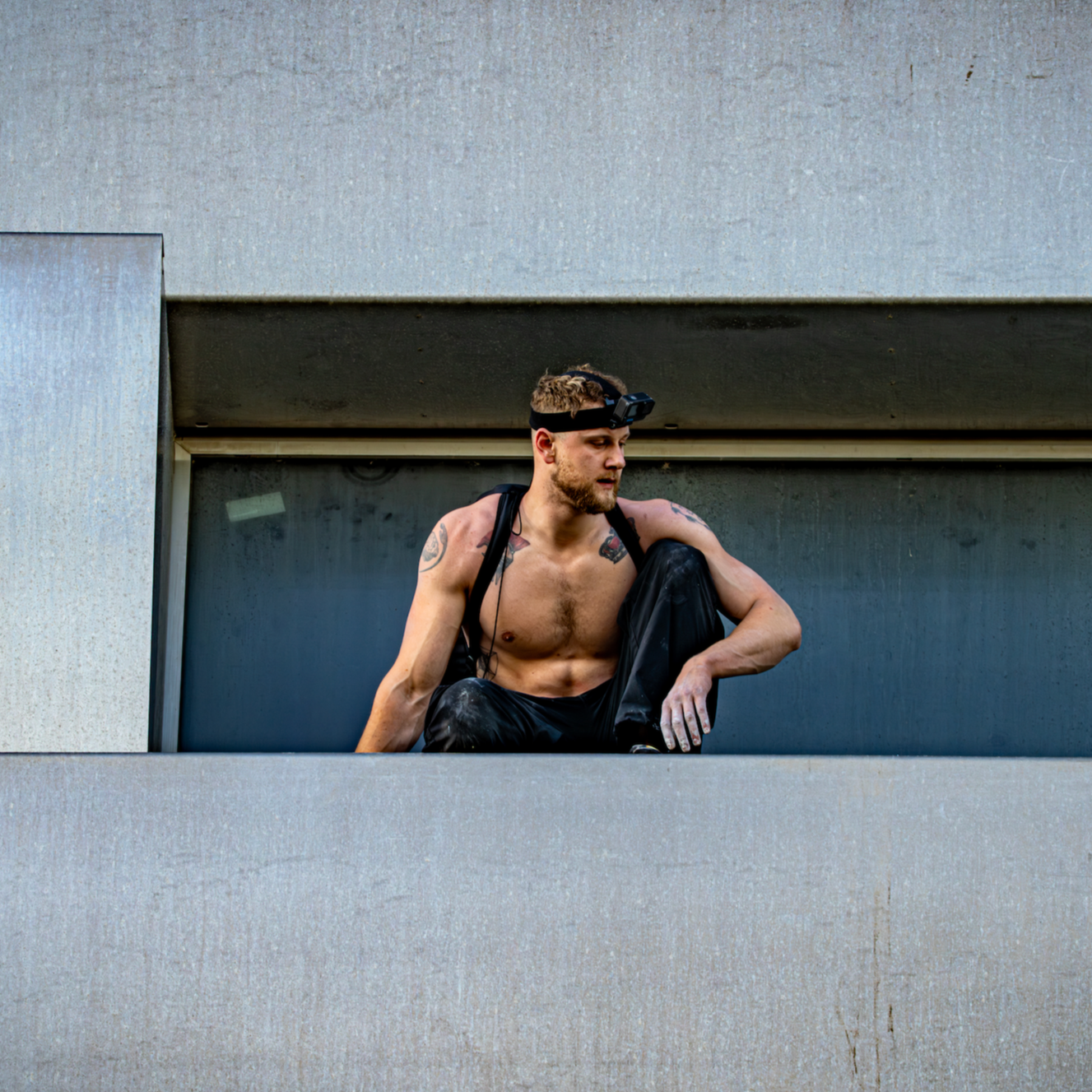 George is sitting on the concrete ledge of a building looking over the side wearing black trousers and a GoPro camera on his head. He has tattoos on his chest and arms.